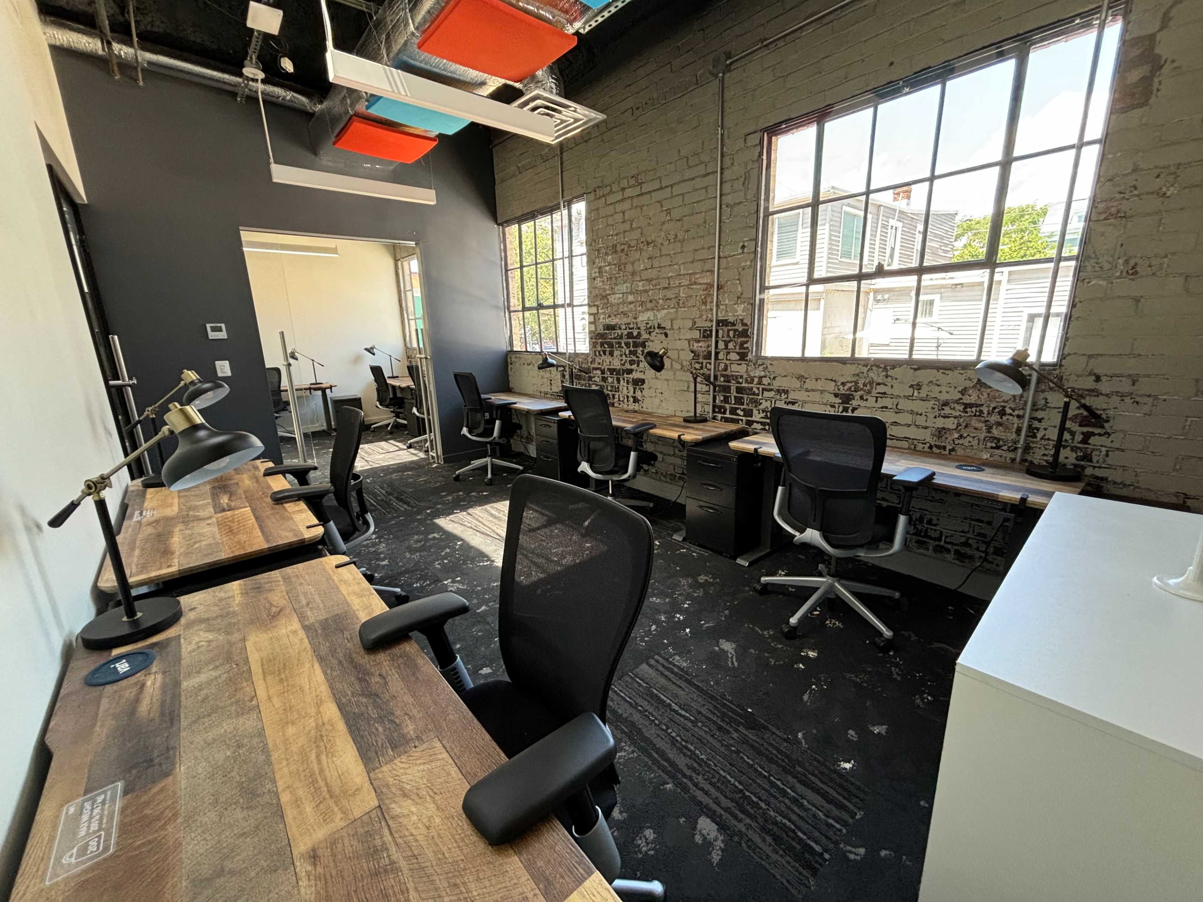 A spacious office with several desks and chairs arranged near large windows, along with exposed brick walls and industrial lighting.