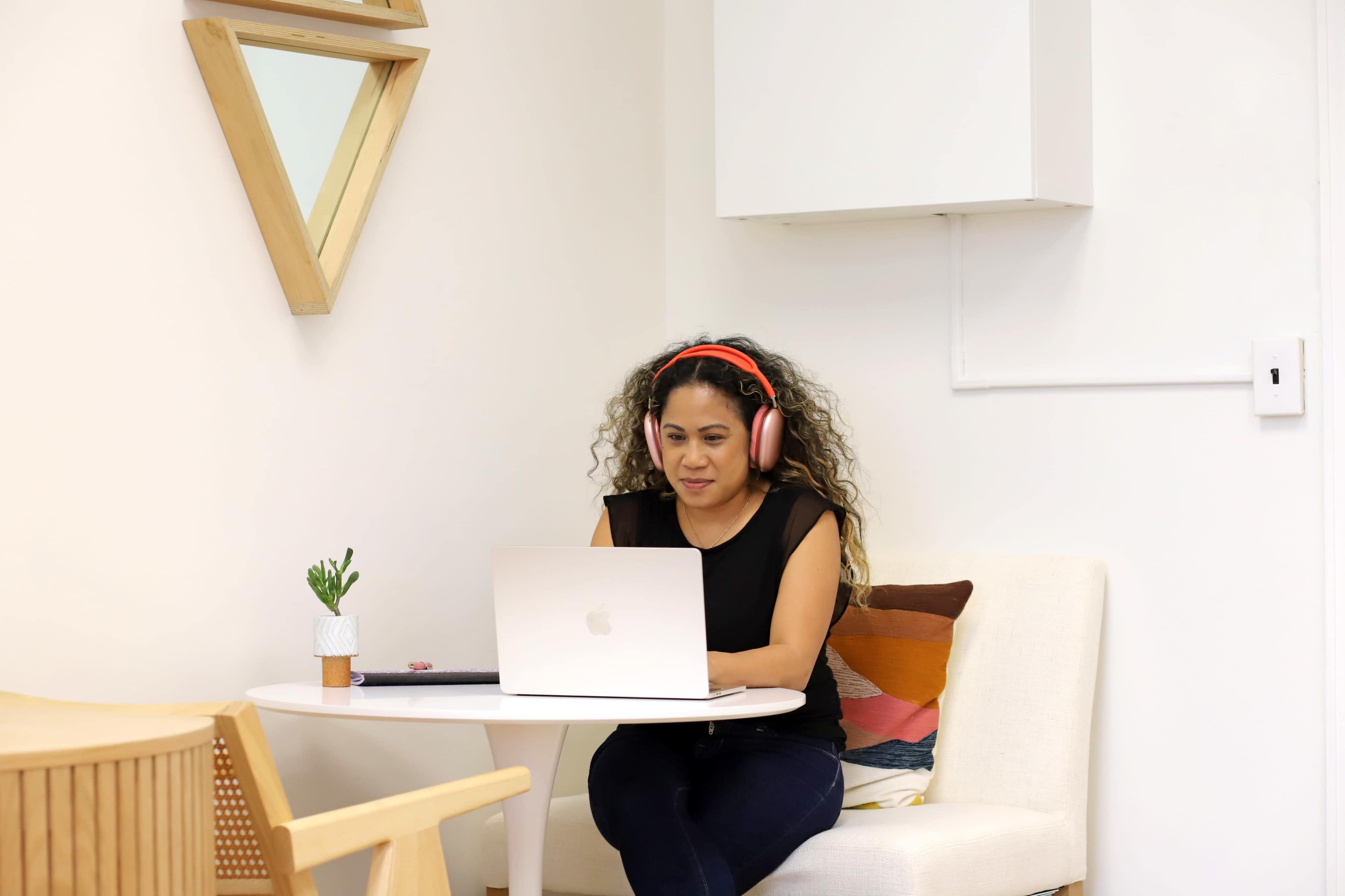 A woman with curly hair, wearing headphones, is sitting at a desk in a minimalist space, working on a laptop.