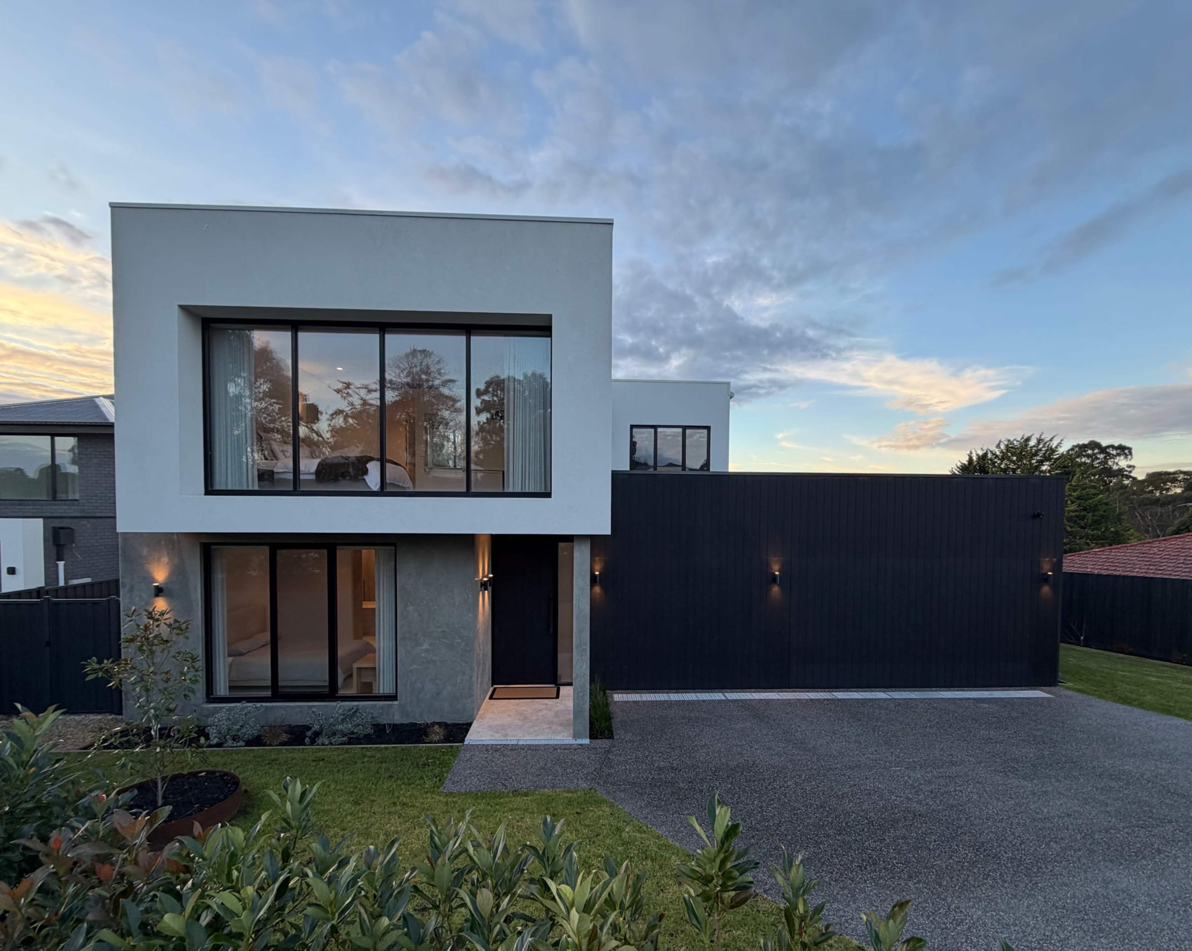 A modern two-story house features large windows and a combination of light and dark exterior materials, set against a backdrop of trees and an evening sky.