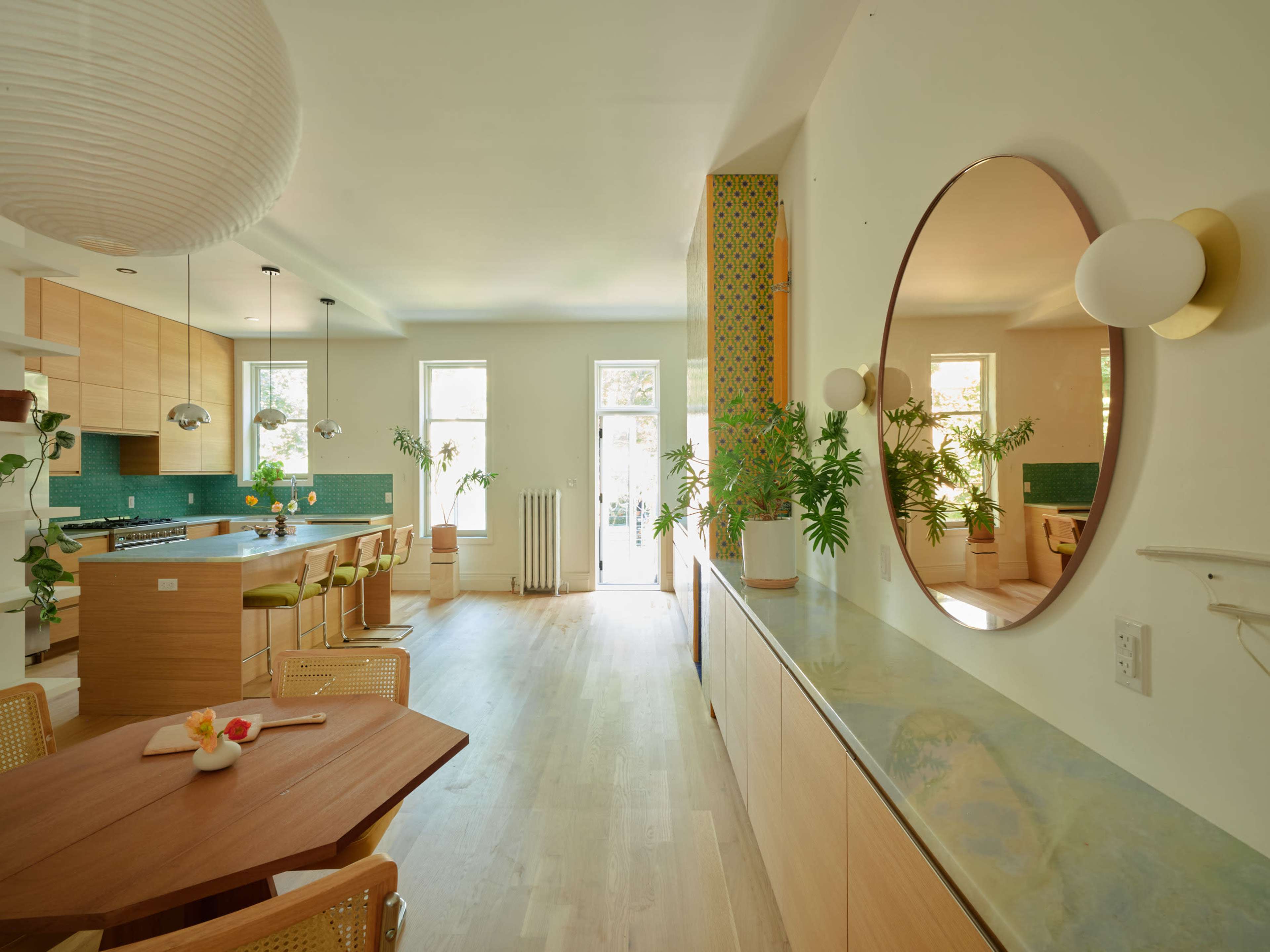 The image depicts a modern kitchen and dining area featuring wooden cabinetry, a large round mirror, and various indoor plants.