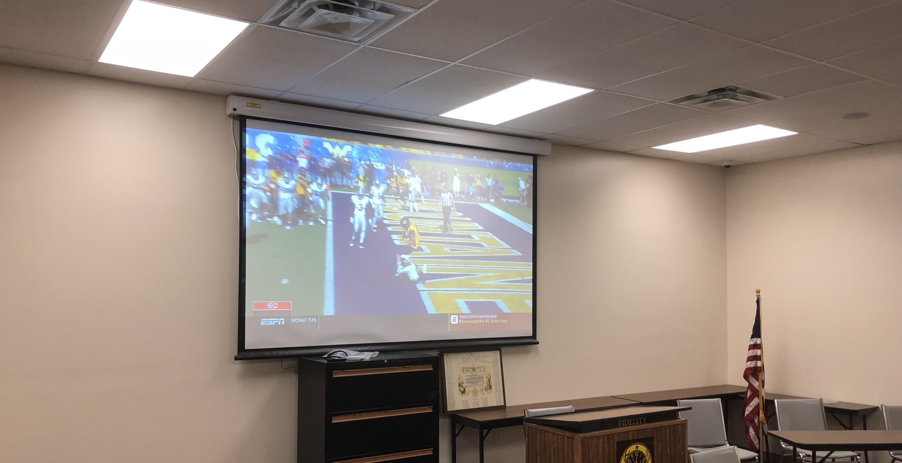 A large screen displays a sports broadcast in a meeting room, with an American flag visible in the background.