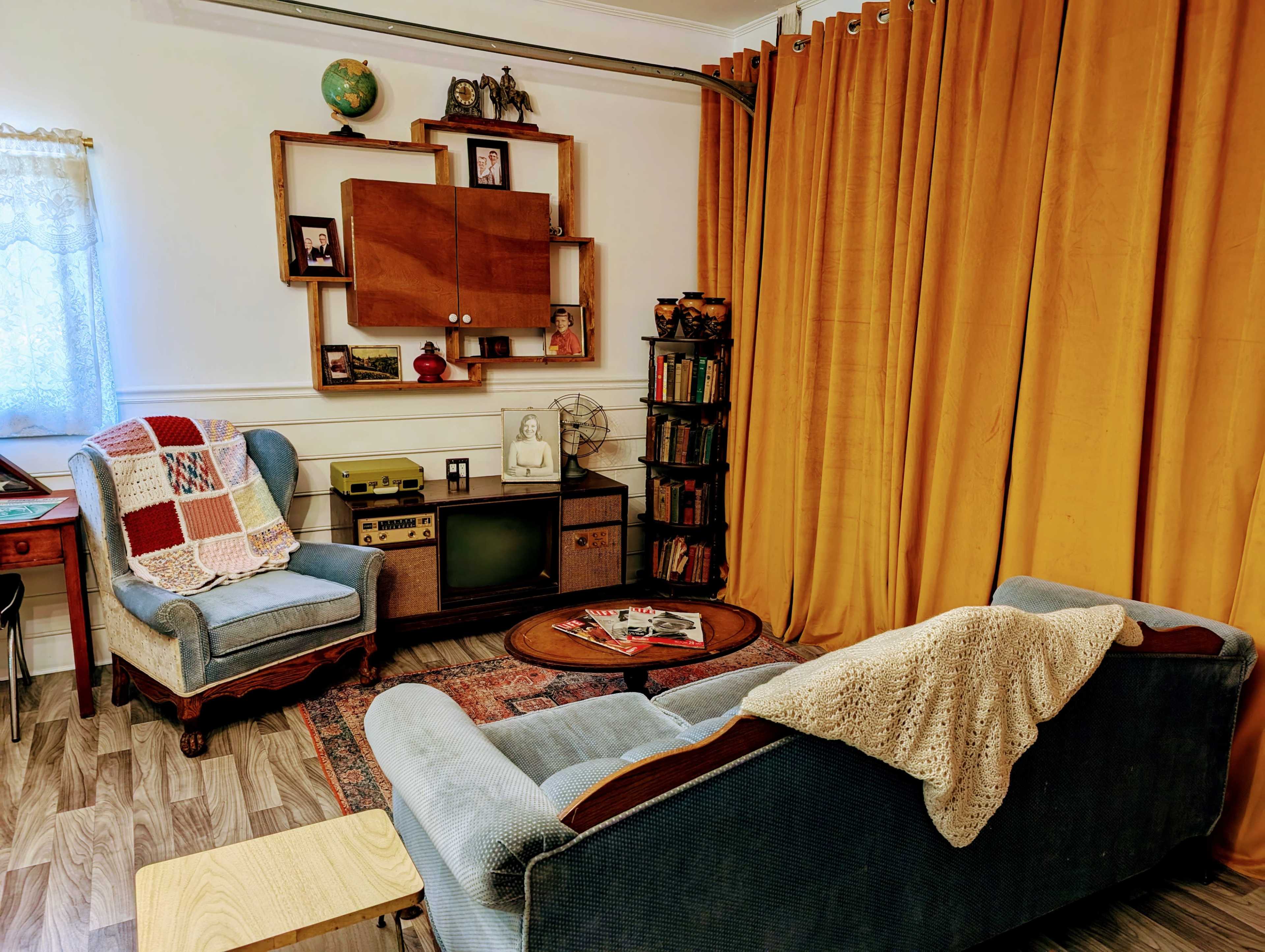 A cozy living room with two upholstered chairs, a wooden coffee table, a vintage television, shelves filled with books, and yellow curtains.