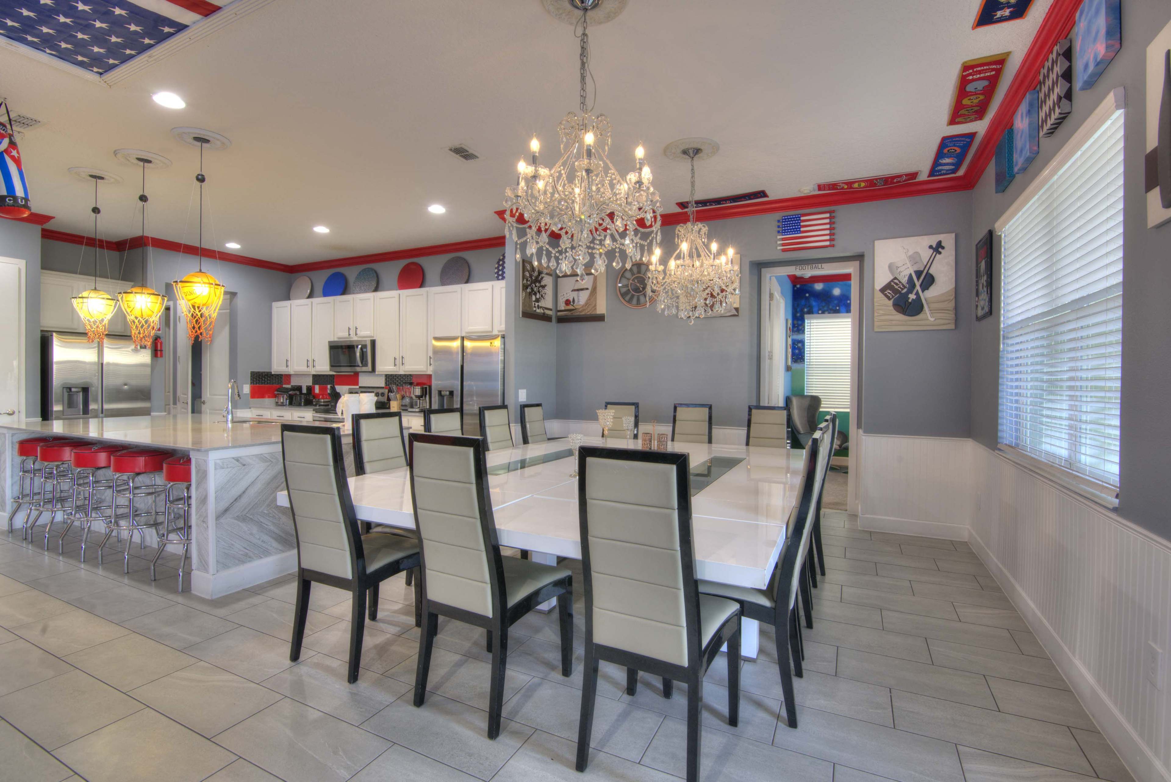 A spacious dining area features a long white table surrounded by black chairs, with modern chandeliers and colorful decorative elements on the ceiling.