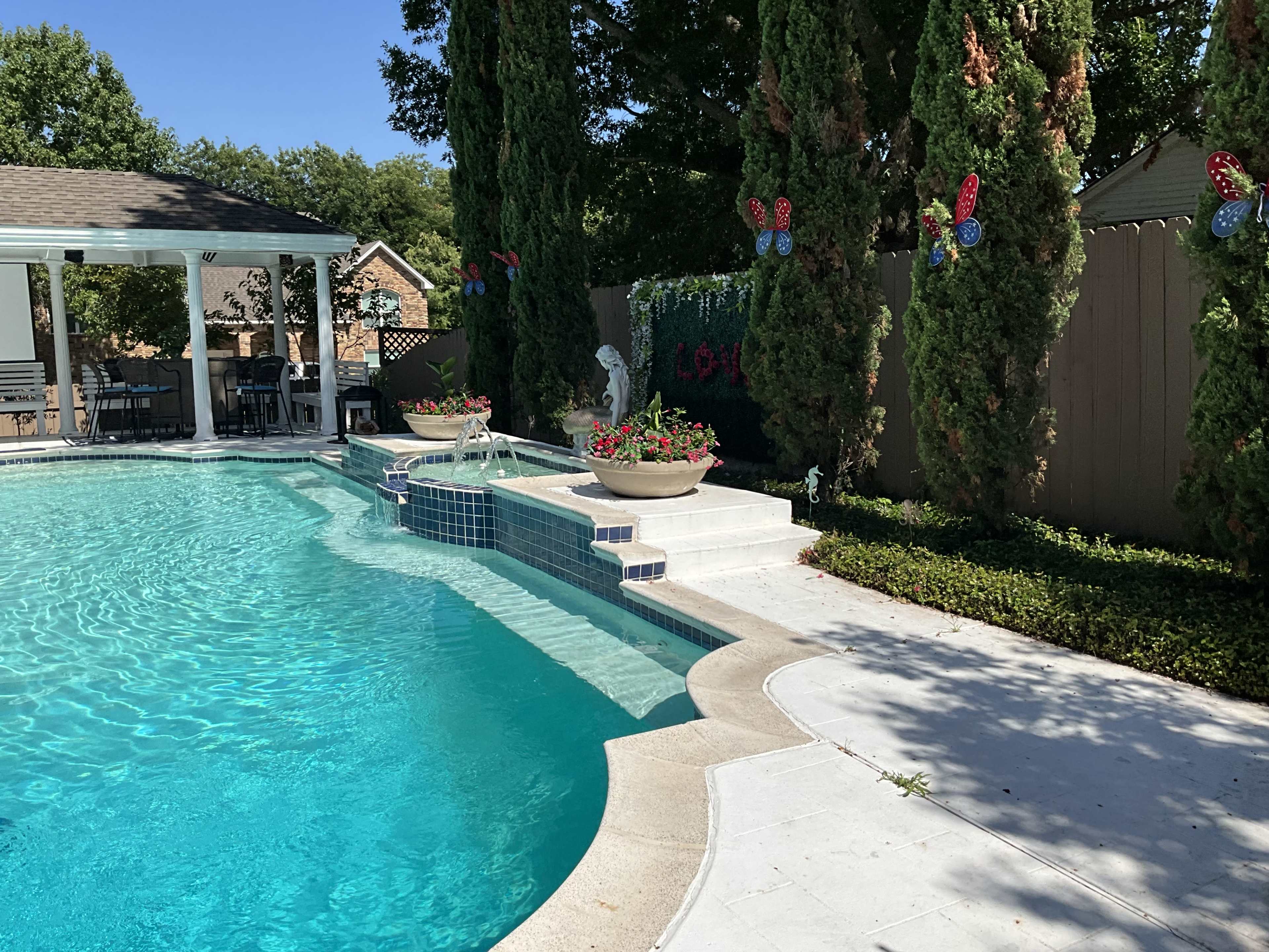 A clear swimming pool with steps leading up to a landscaped area featuring potted flowers and decorative butterflies along a fence.