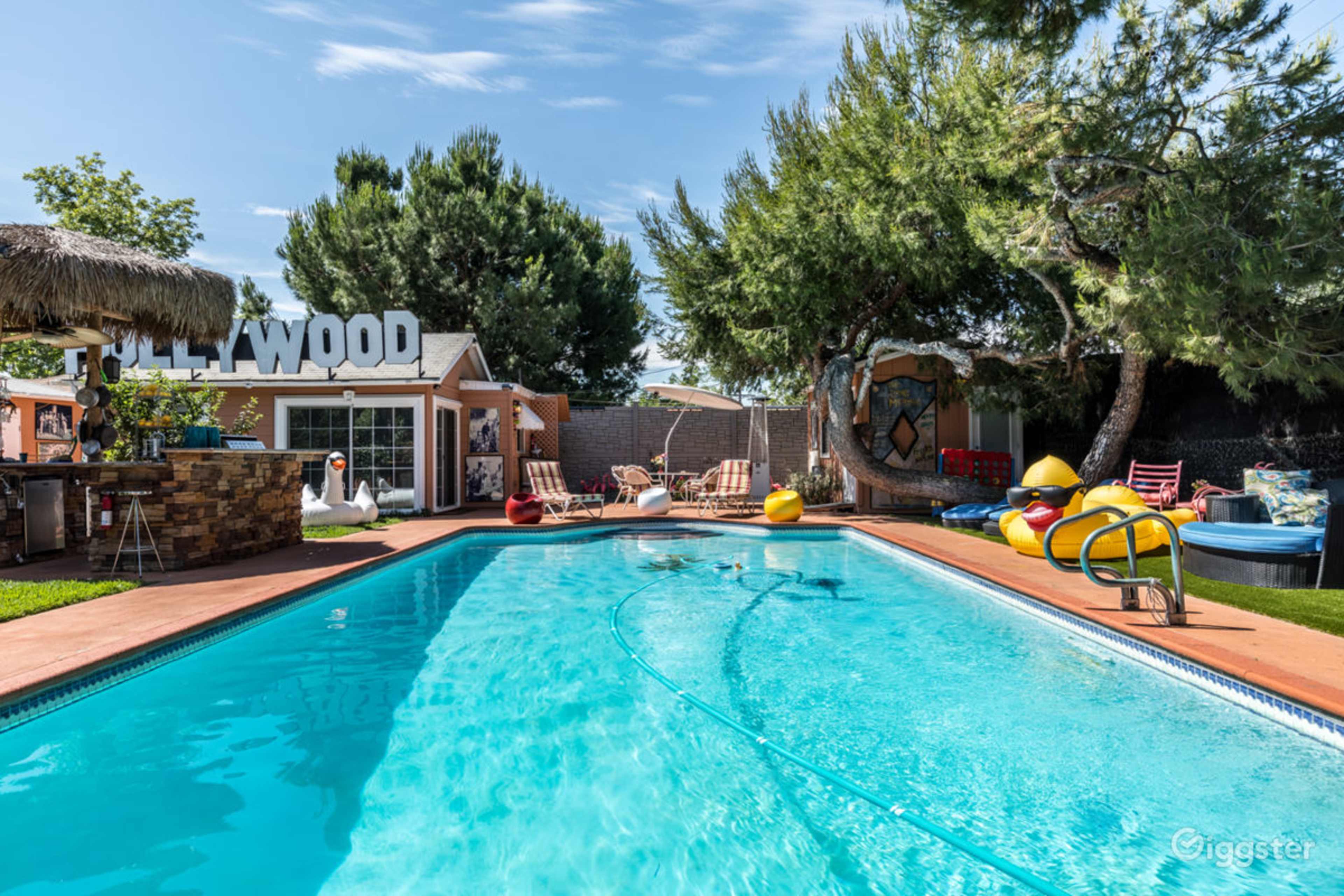 A vibrant pool area with a clear blue pool, colorful lounge chairs, and a "HOLLYWOOD" sign in the background surrounded by trees and a tiki-style hut.