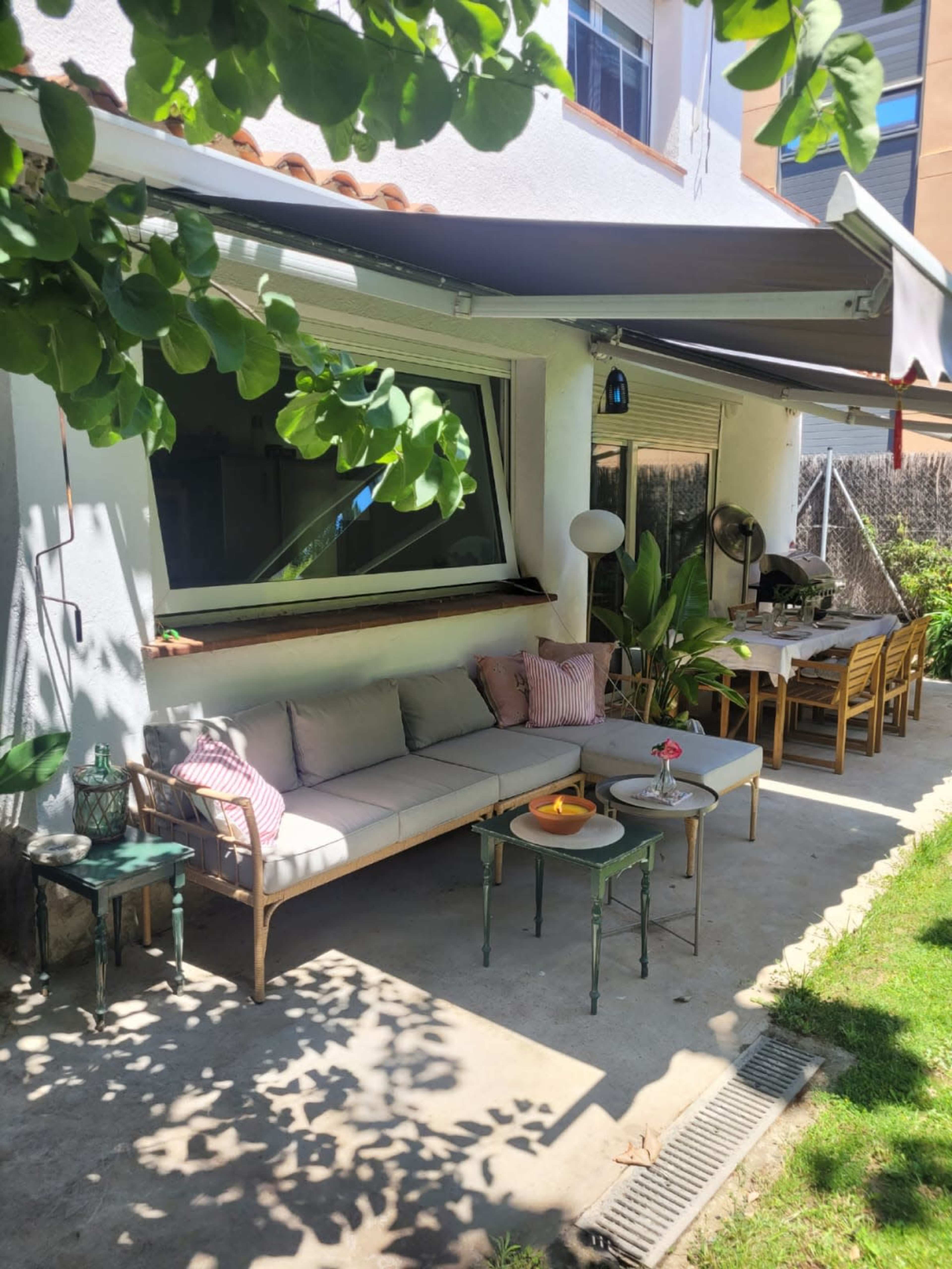 The image shows a cozy outdoor seating area with a sofa, small tables, and a dining table underneath a shaded patio.