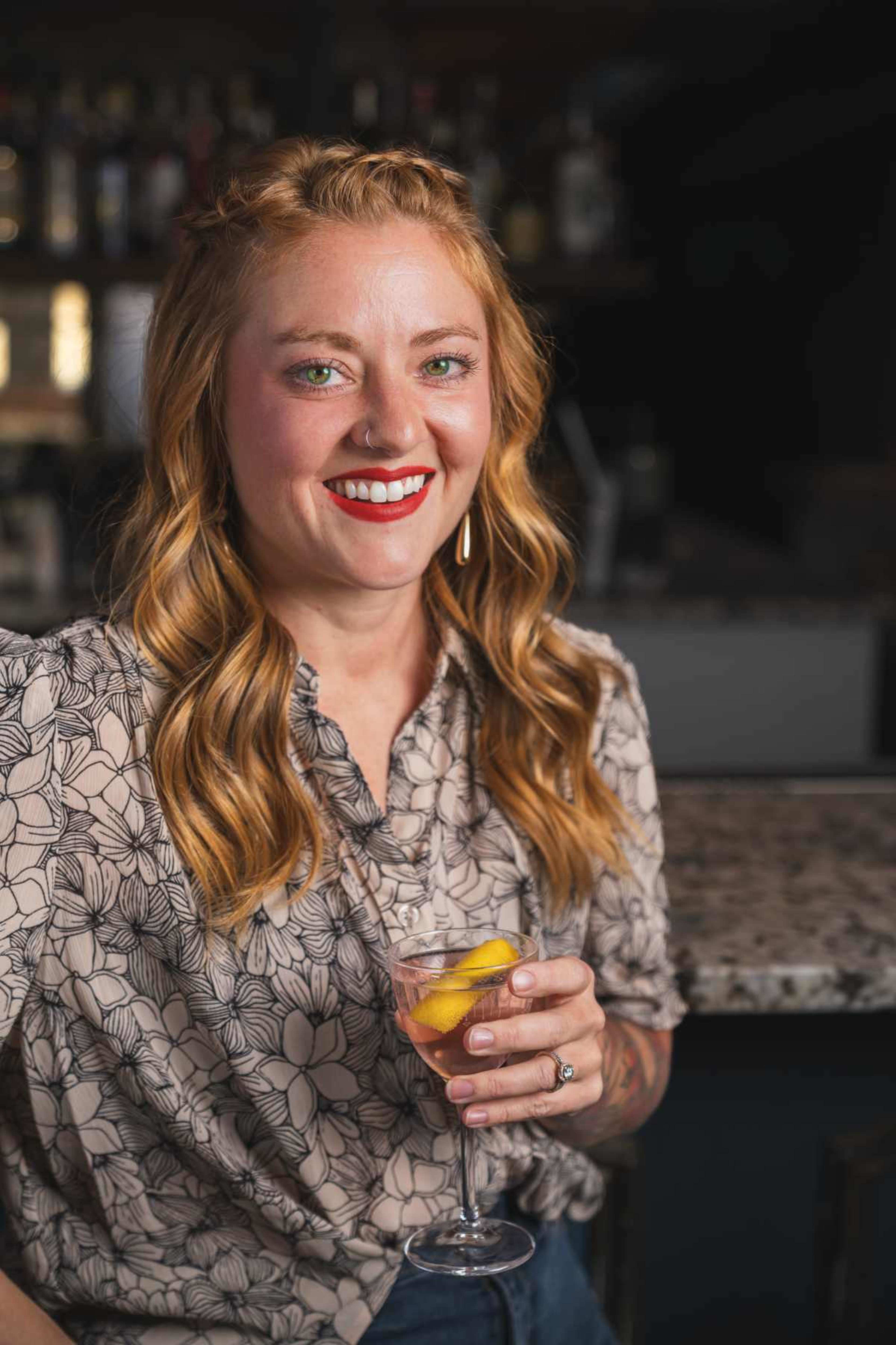 A woman with wavy, shoulder-length hair smiles while holding a glass of cocktail with lemon slices, seated at a bar.