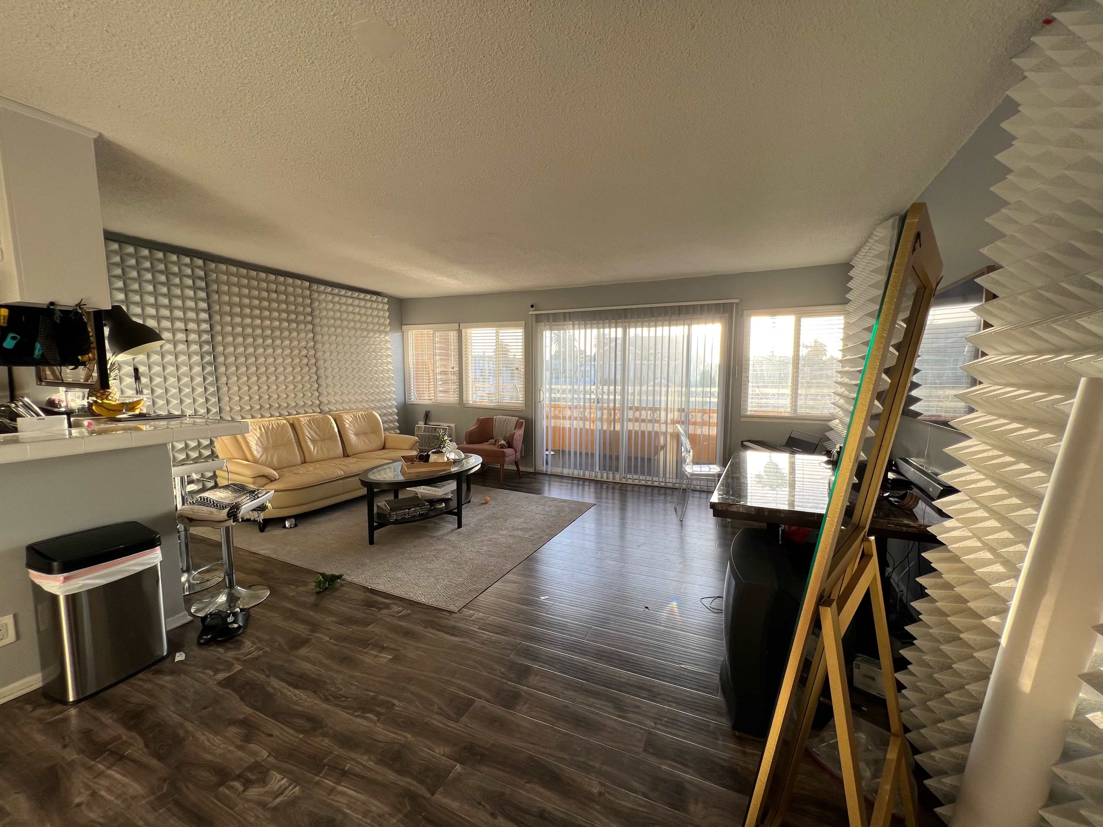 A modern living room with a beige sofa, a glass coffee table, and large windows covered by sheer curtains, set in a space with patterned wall panels.