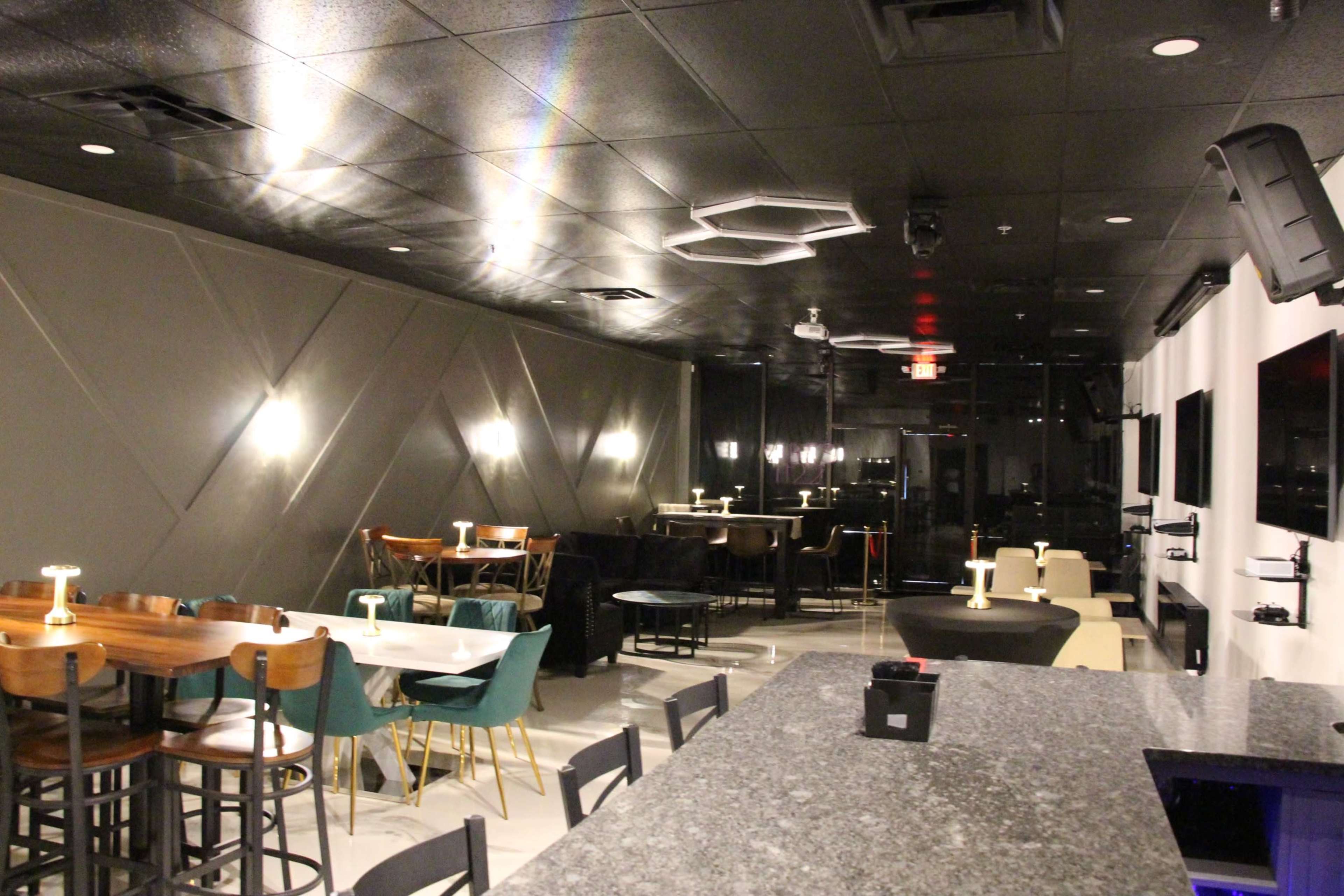 A modern, sparsely furnished restaurant interior featuring wooden and metal tables, chairs, and a bar counter, illuminated by ambient lighting.