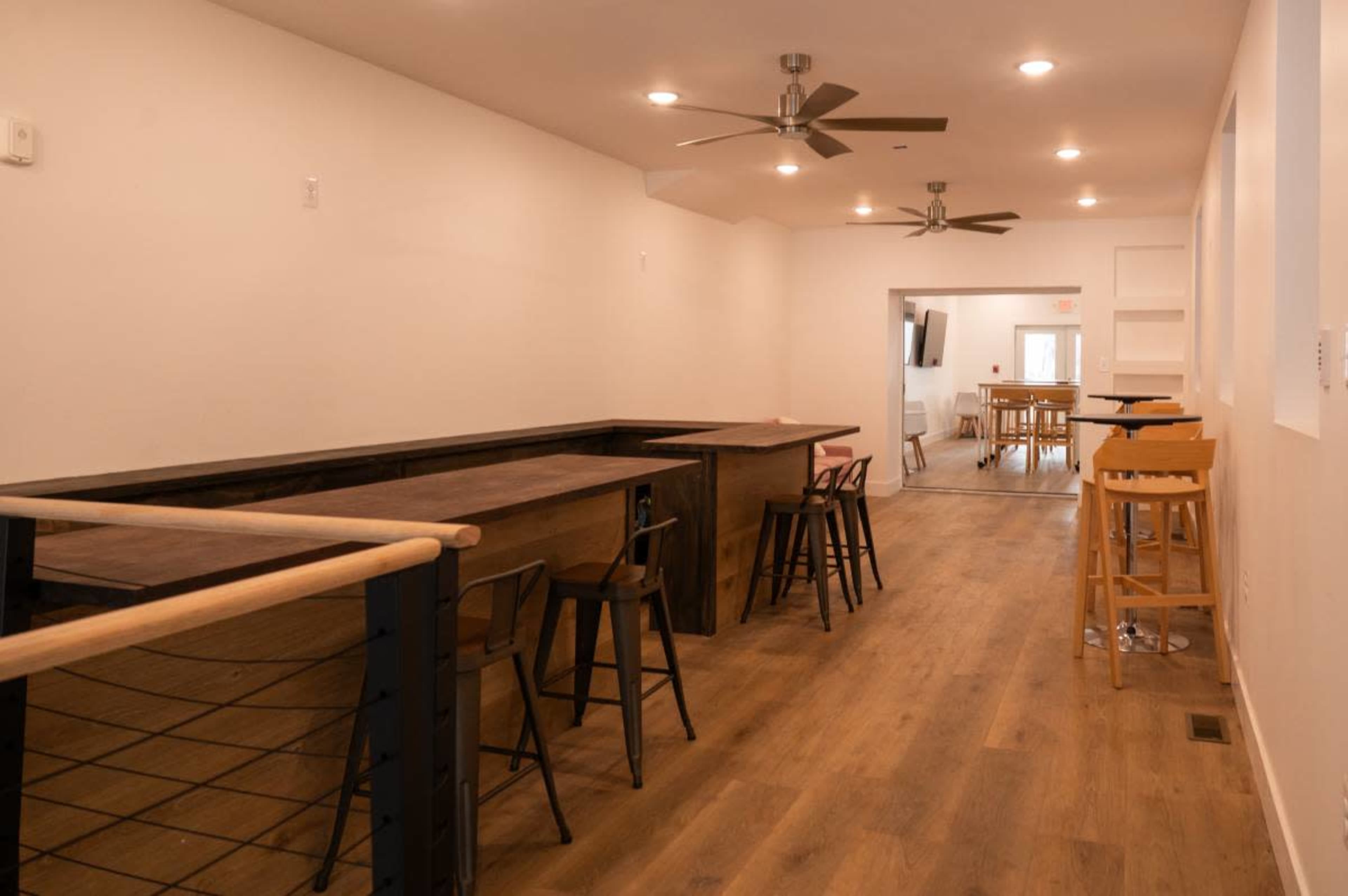 The image shows a modern, minimally furnished interior space with bar-height seating and wooden floors, featuring ceiling fans and a light color palette.