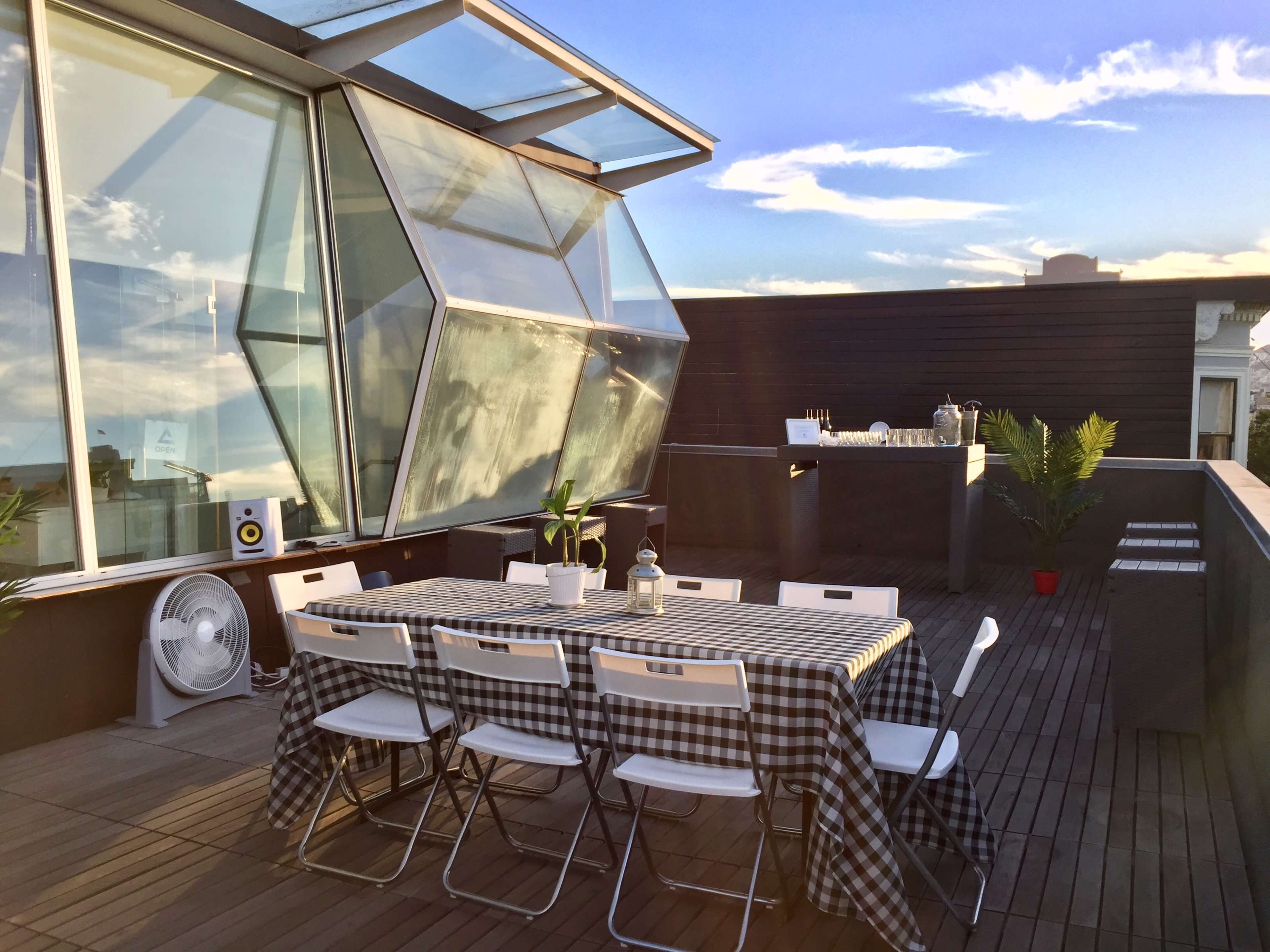 A modern rooftop terrace features a large table set with a checkered tablecloth, surrounded by folding chairs, and a glass structure in the background.