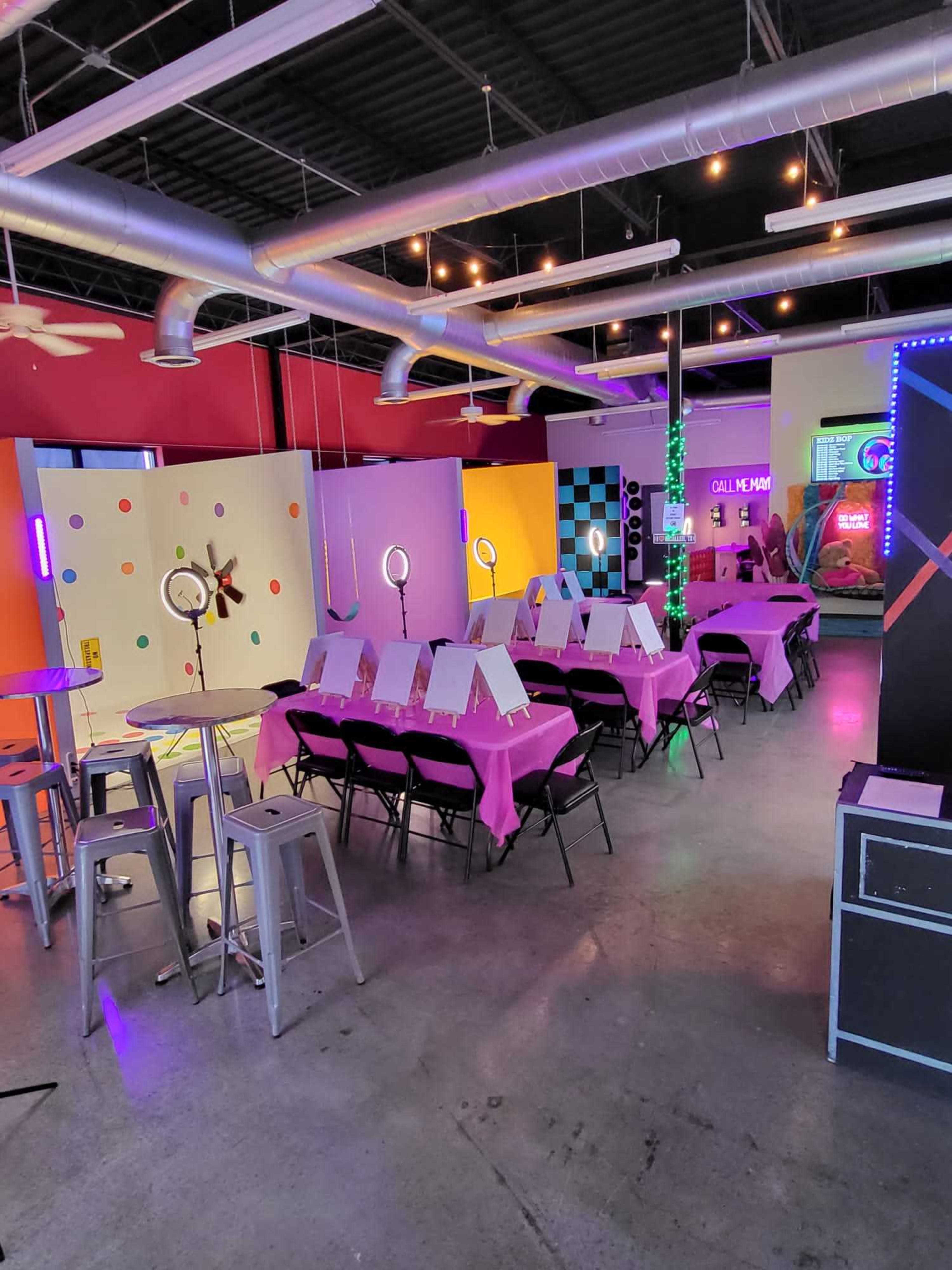 A brightly colored event space set up with tables covered in pink cloths, each featuring easels with blank canvases, alongside various decorative lights and art-related displays.