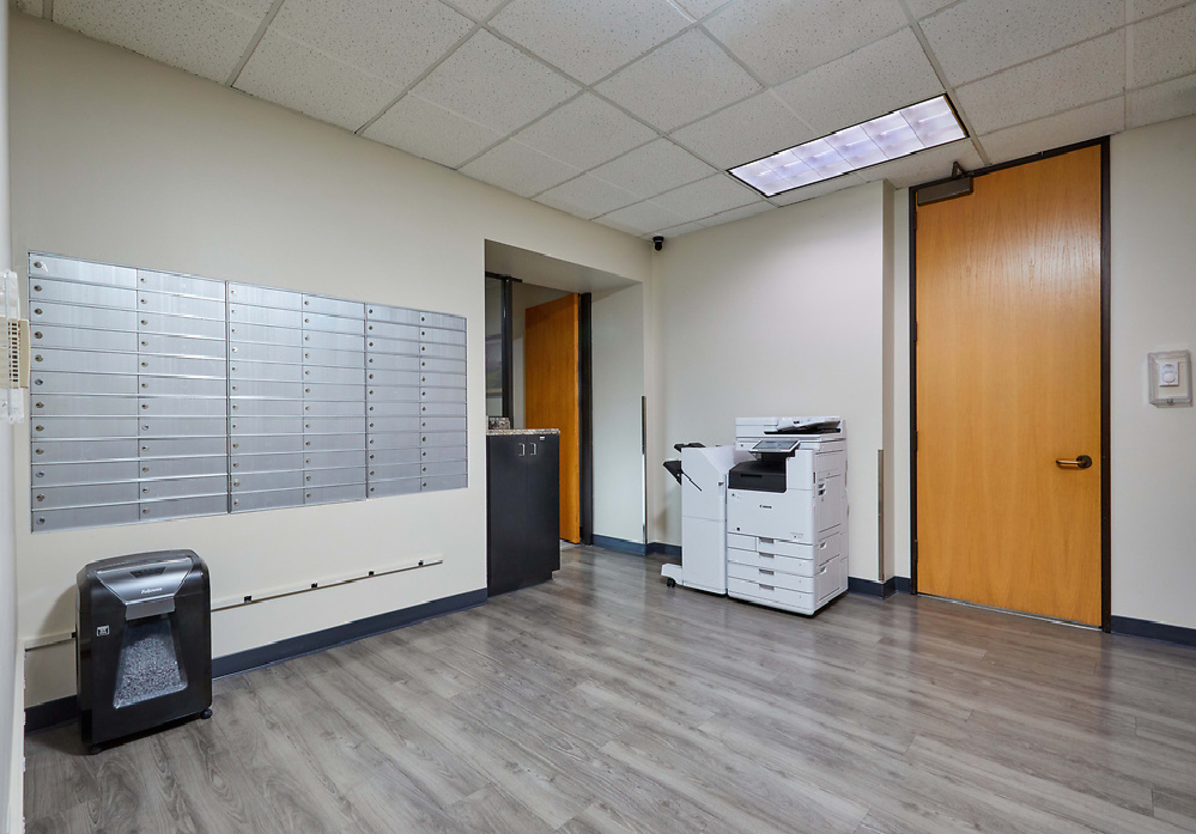 The image shows a corridor with a row of mailboxes on the left, a copier on the right, and a wooden door at the end.