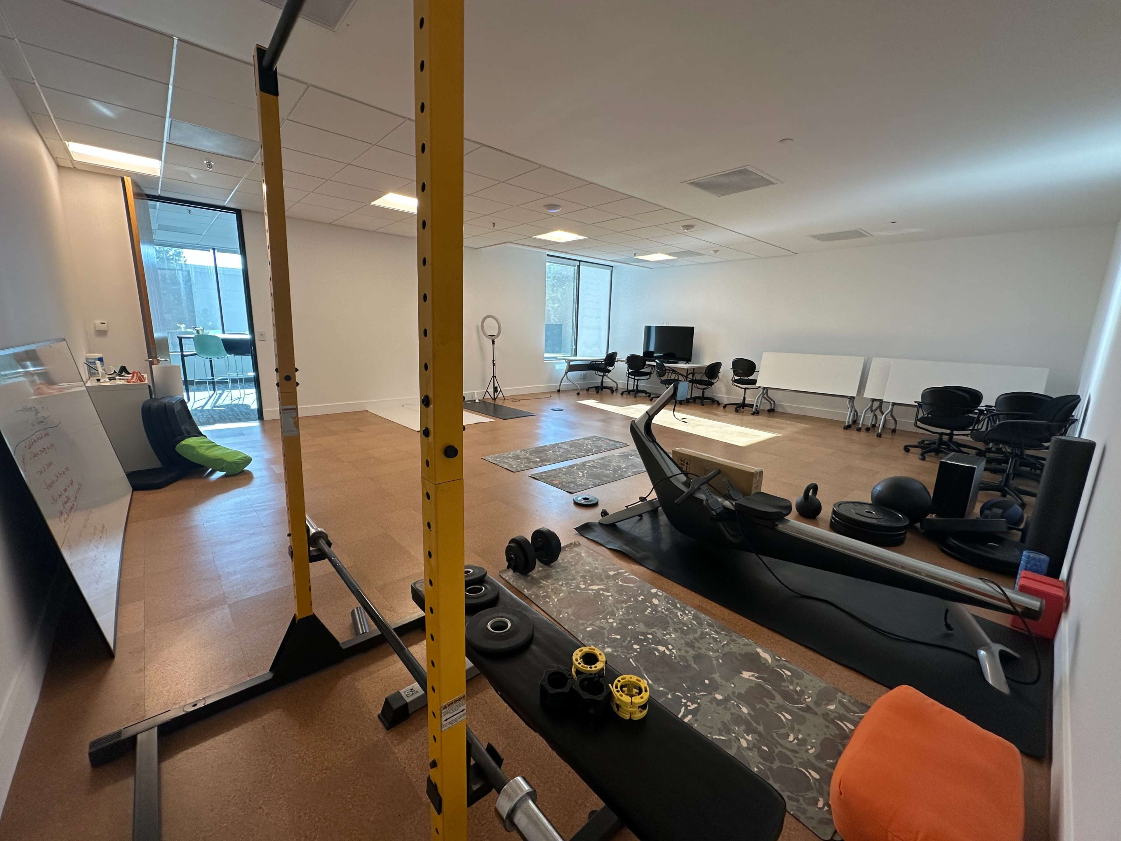 The image shows an empty gym or workout space equipped with exercise equipment, including a squat rack, weights, and various fitness mats, alongside office furniture like chairs and tables.