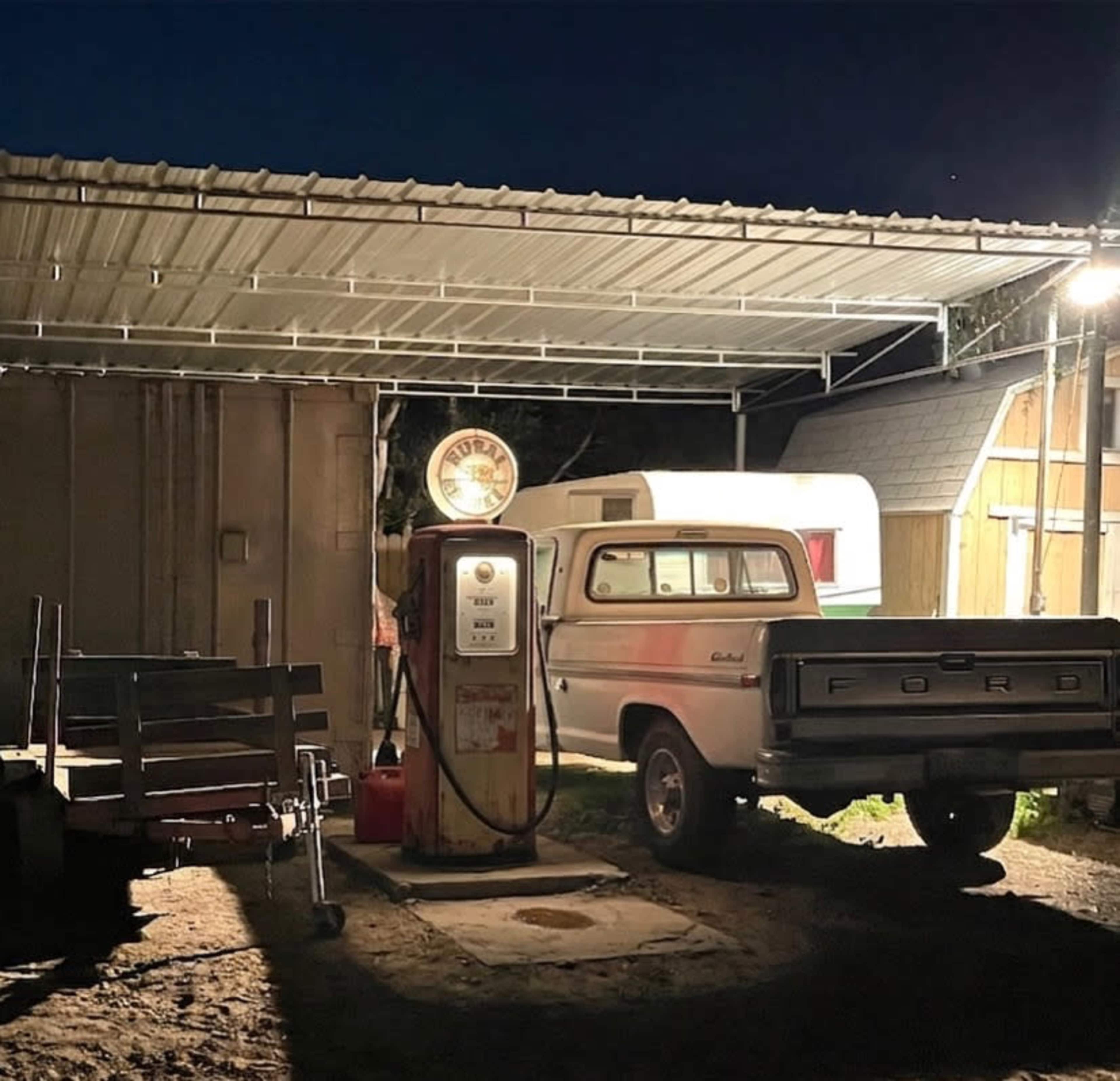 The image depicts a vintage gas pump next to a classic Ford truck and a parked caravan under a lit canopy at night.