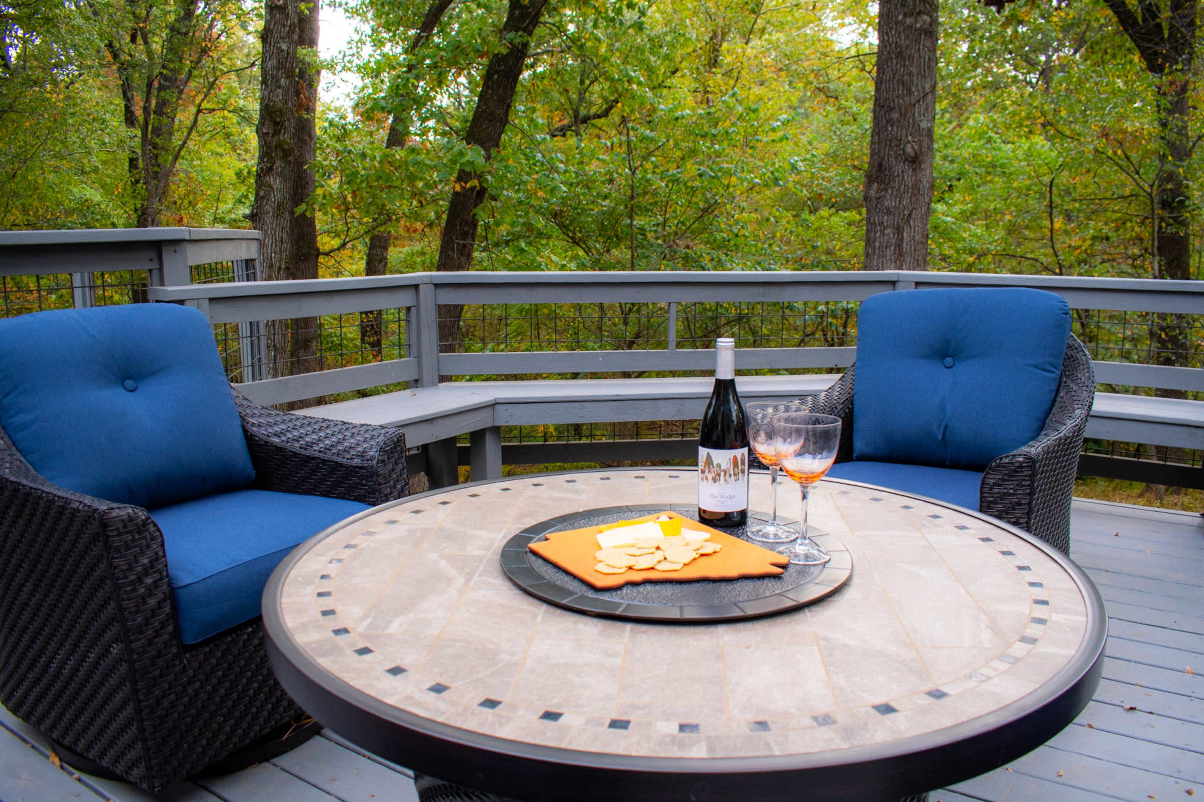 A patio setting features two blue cushioned chairs, a round table with snacks, and a bottle of wine beside two glasses against a backdrop of trees.