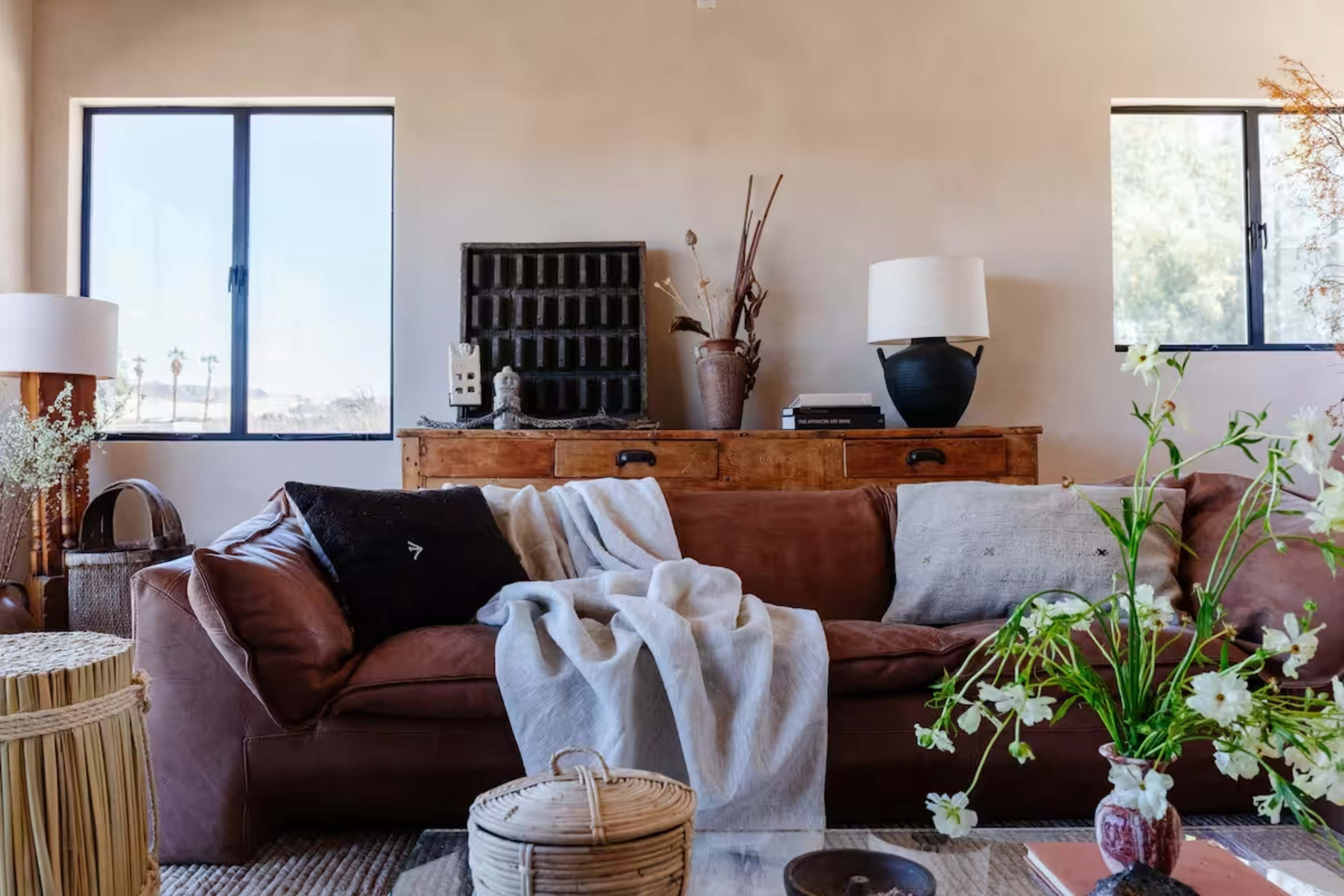 The image shows a cozy living room featuring a brown leather sofa draped with a gray blanket, a wooden cabinet, and decorative plants by large windows.