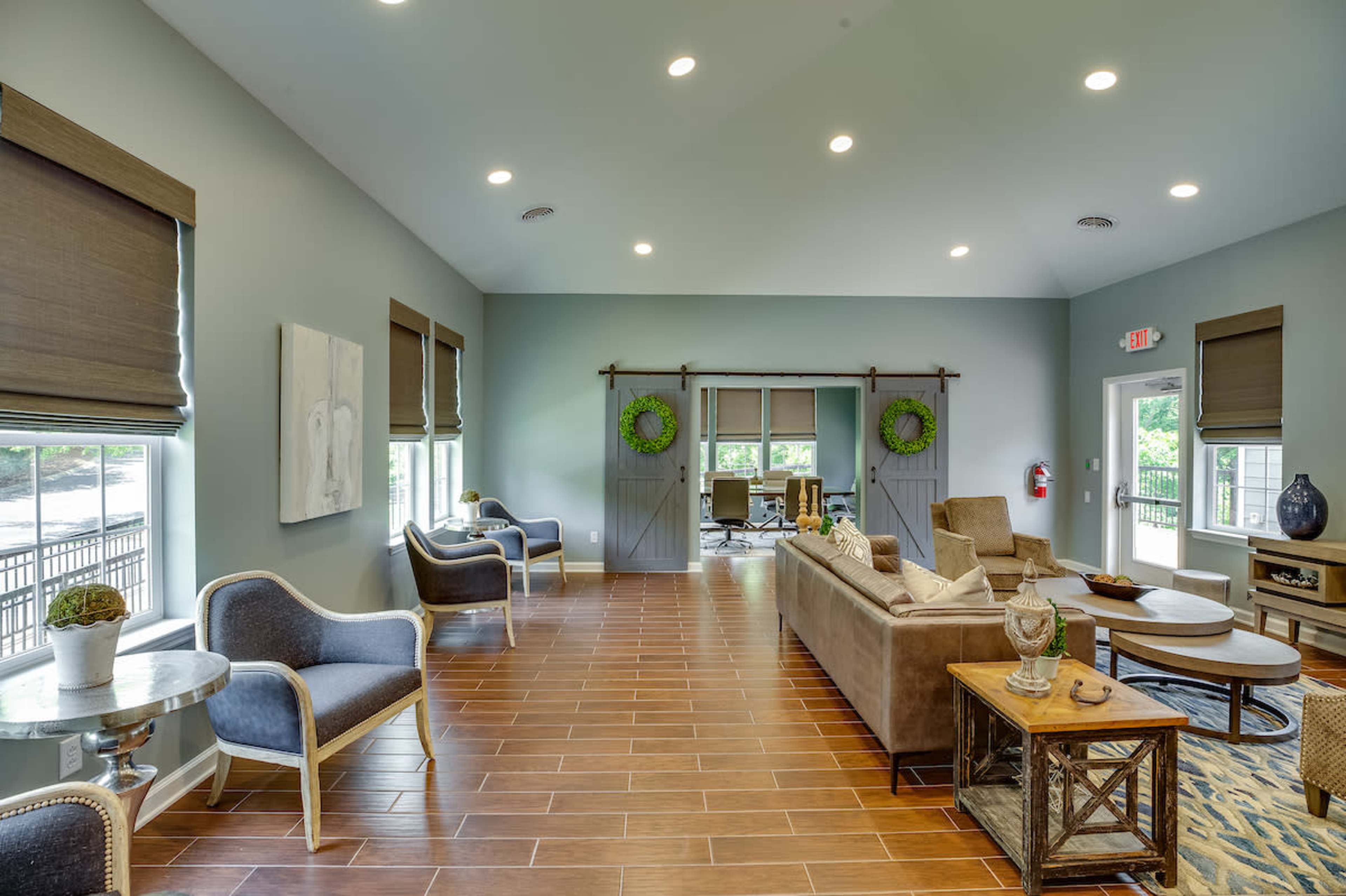 The image shows a well-lit lounge area featuring beige furniture, wooden flooring, and large windows with blinds, creating a cozy atmosphere.