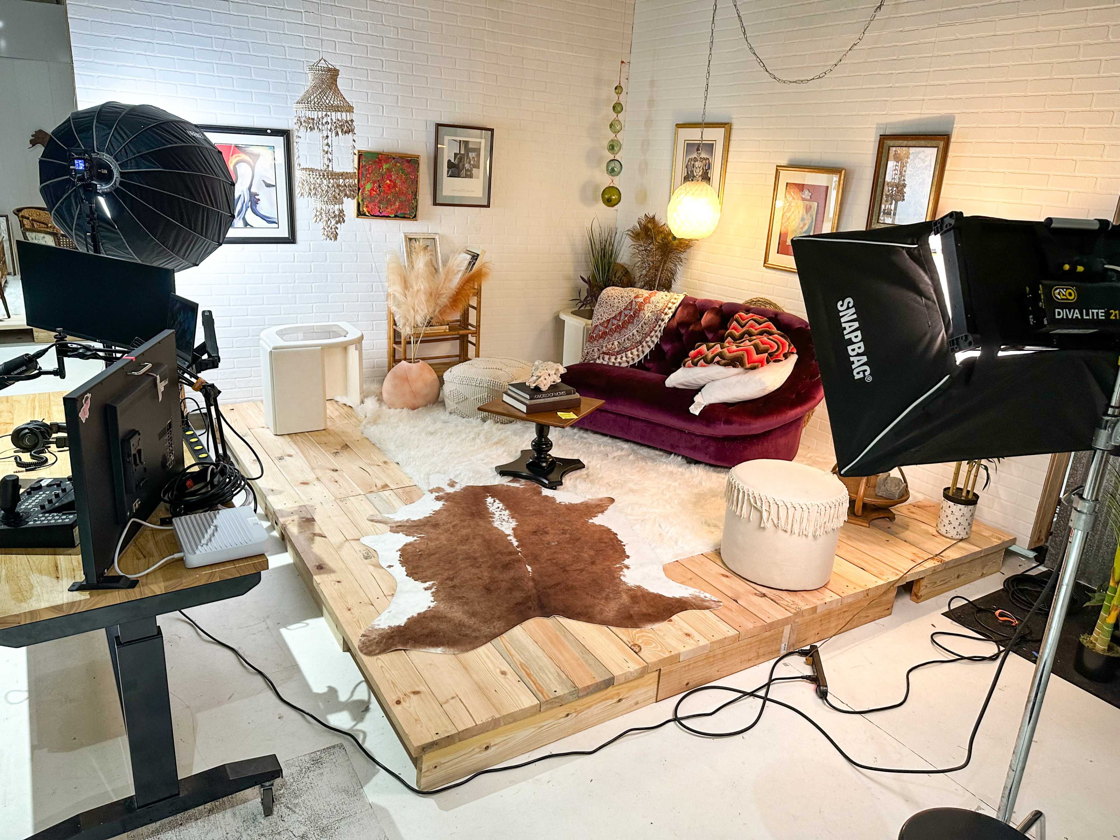 A cozy studio setup featuring a wooden platform with a cowhide rug, a burgundy sofa, and various decorative items, including a large soft light and workstations.