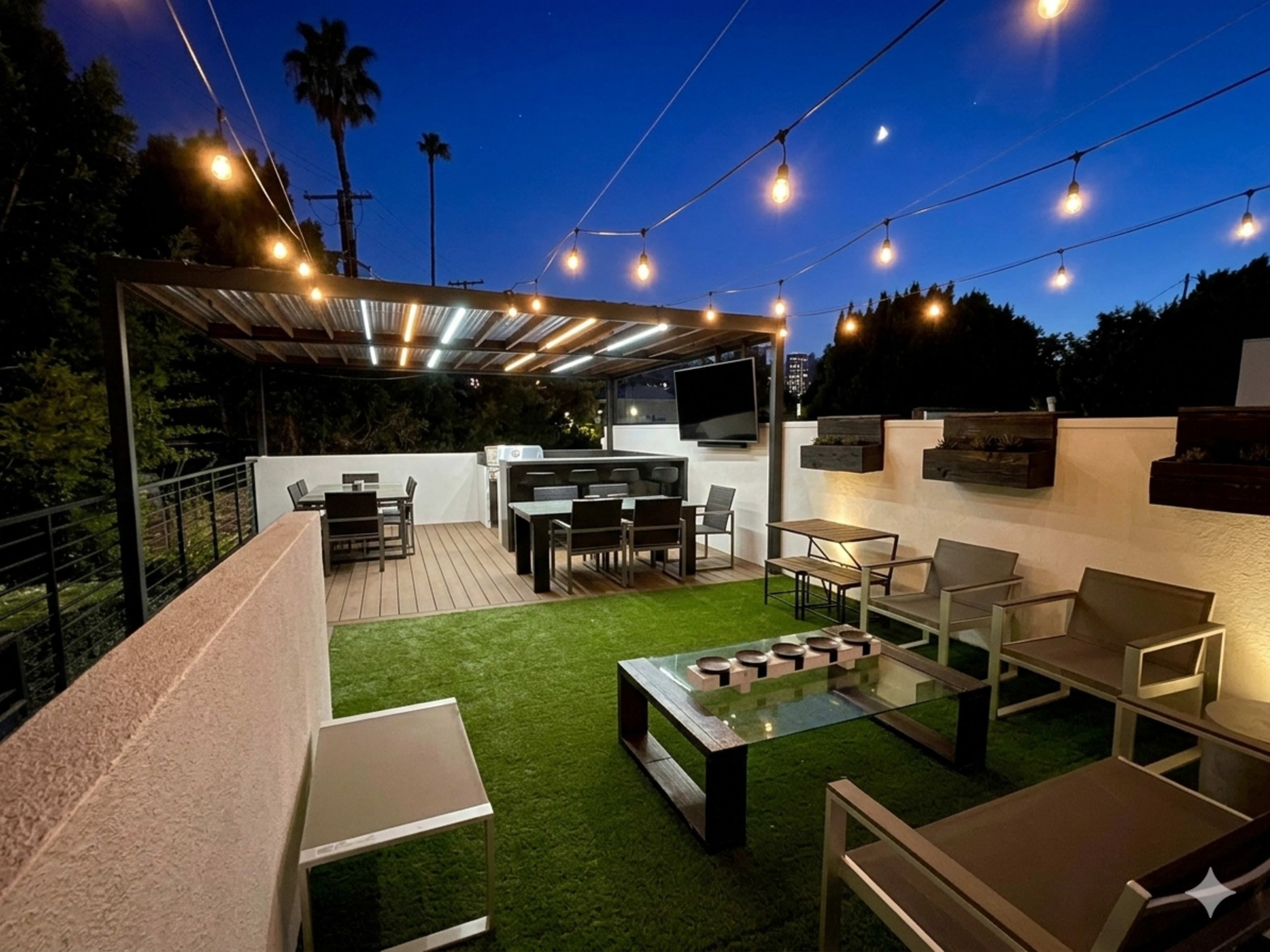 The image shows a rooftop patio at night, featuring outdoor seating, a dining area with tables and chairs, and string lights overhead.