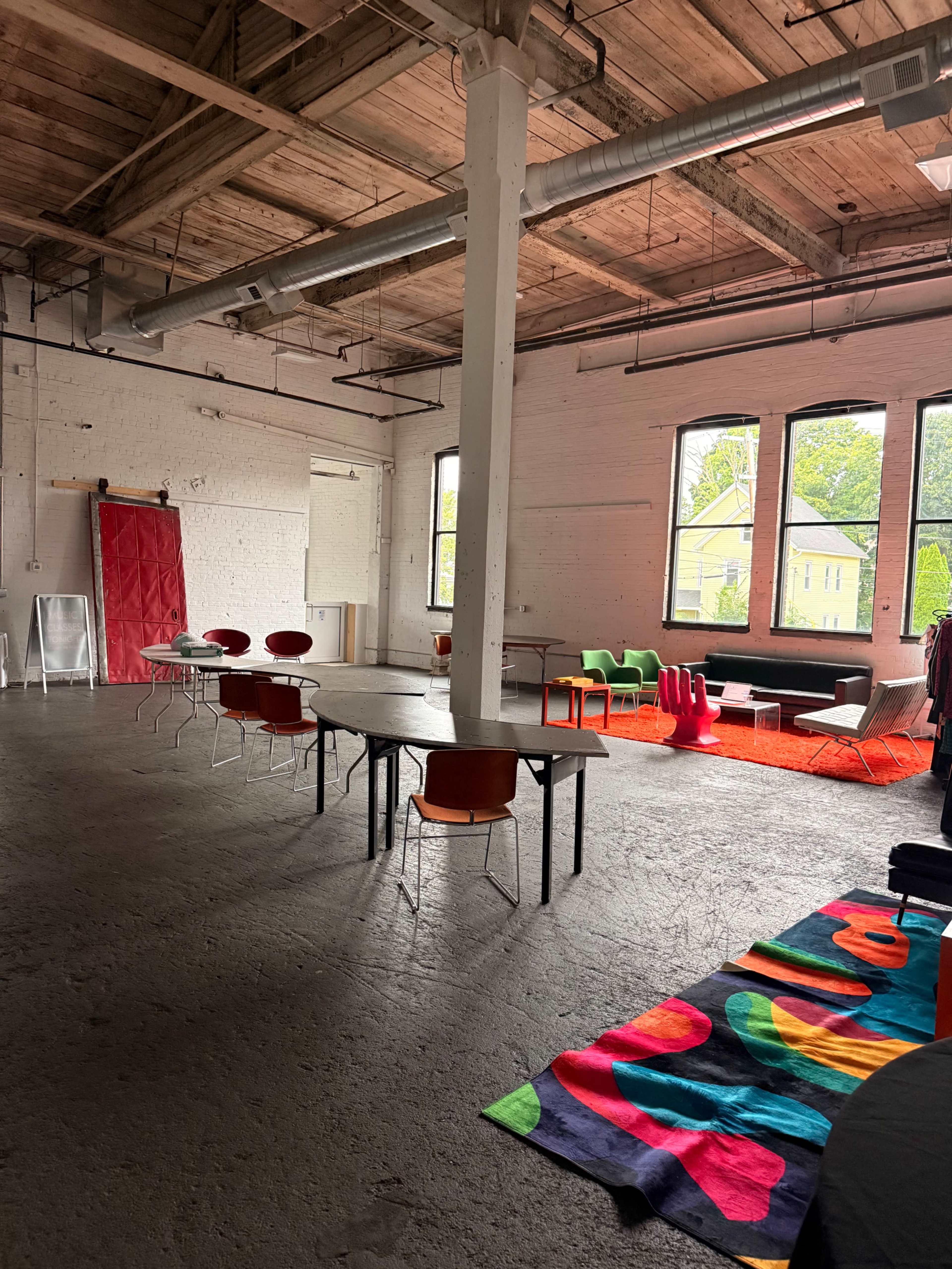 The image shows an industrial-style room featuring a long table with chairs, colorful seating options, and large windows allowing natural light to enter.