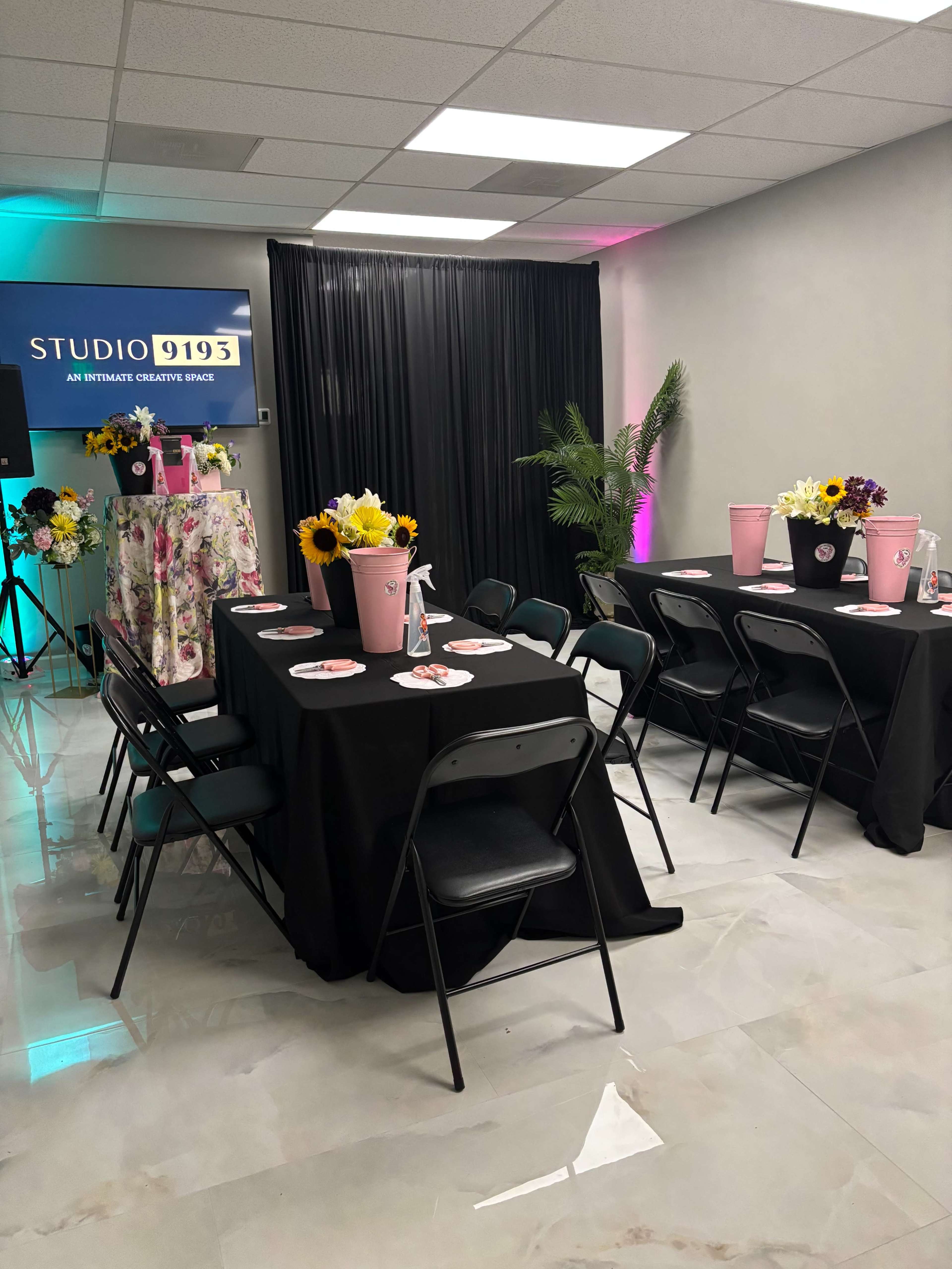 The image shows a decorated indoor event space with black tables set for dining, floral centerpieces, and stylish lighting in the background.
