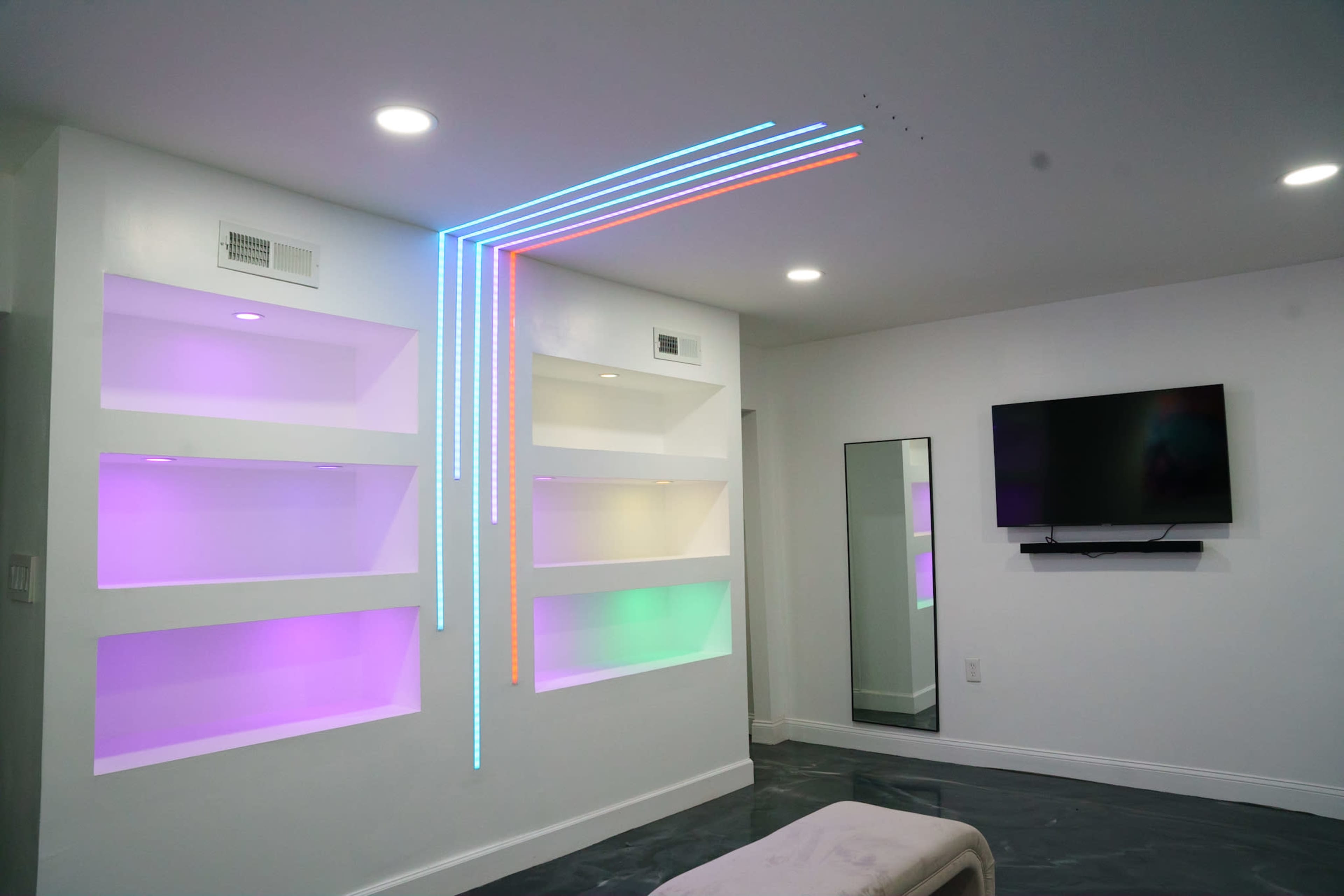 A modern living room with white walls, illuminated shelves in various colors, and linear LED lights along one wall.