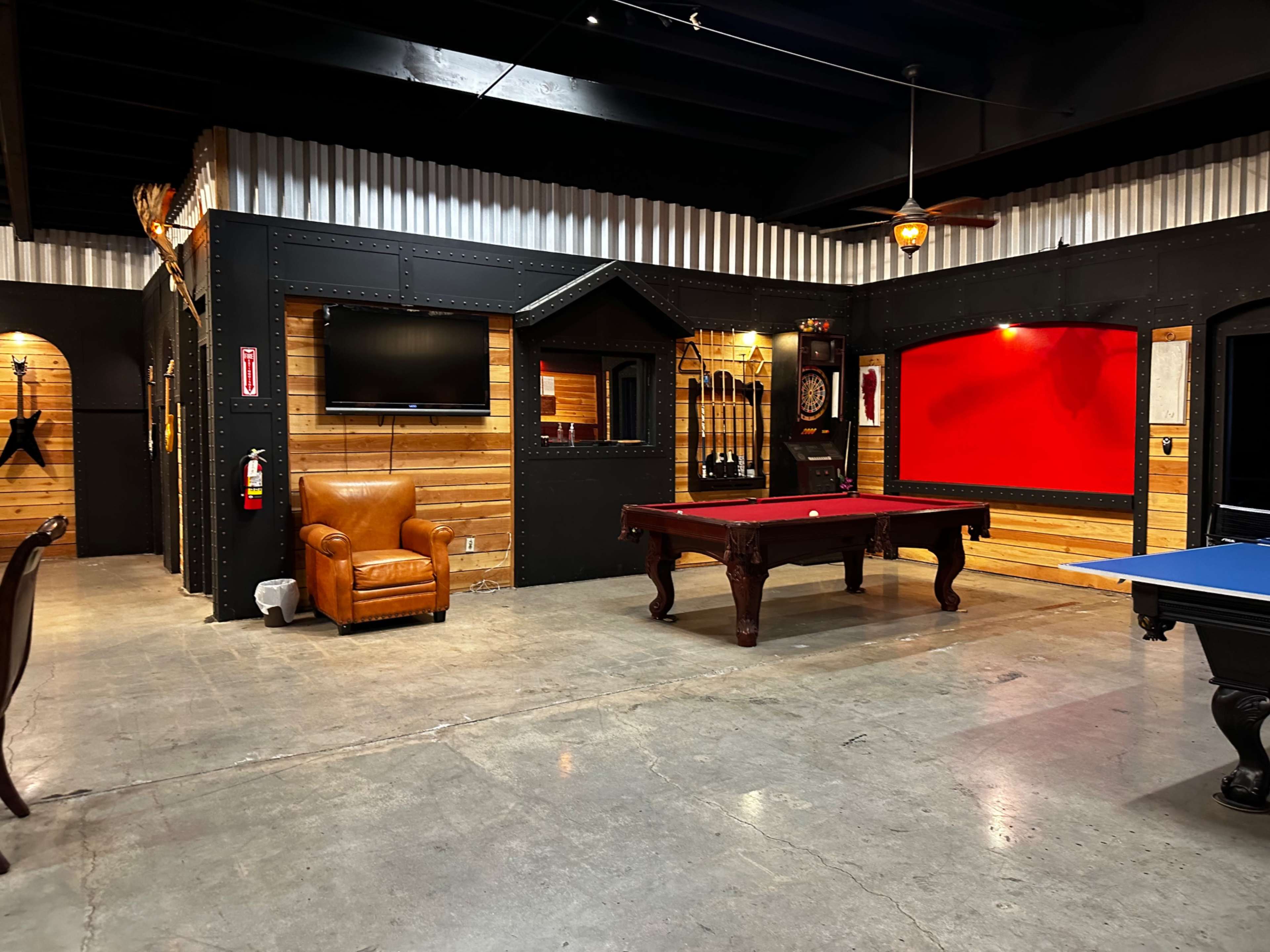 A game room featuring a pool table, a blue ping pong table, a television on the wall, and leather seating in a rustic setting with wooden and metal accents.