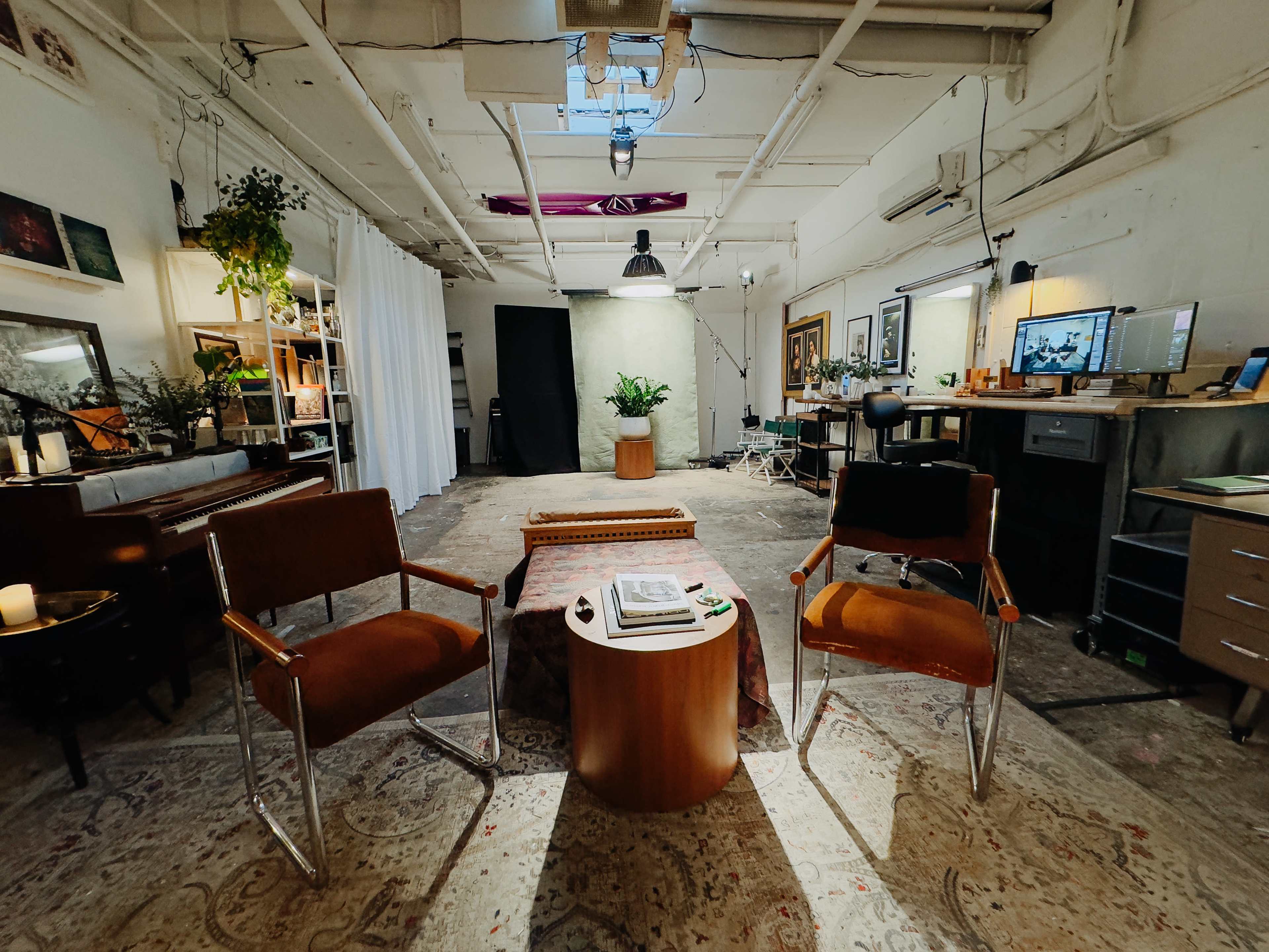 The image shows a spacious studio-like interior with two chairs facing a low table, surrounded by various plants and workspaces.