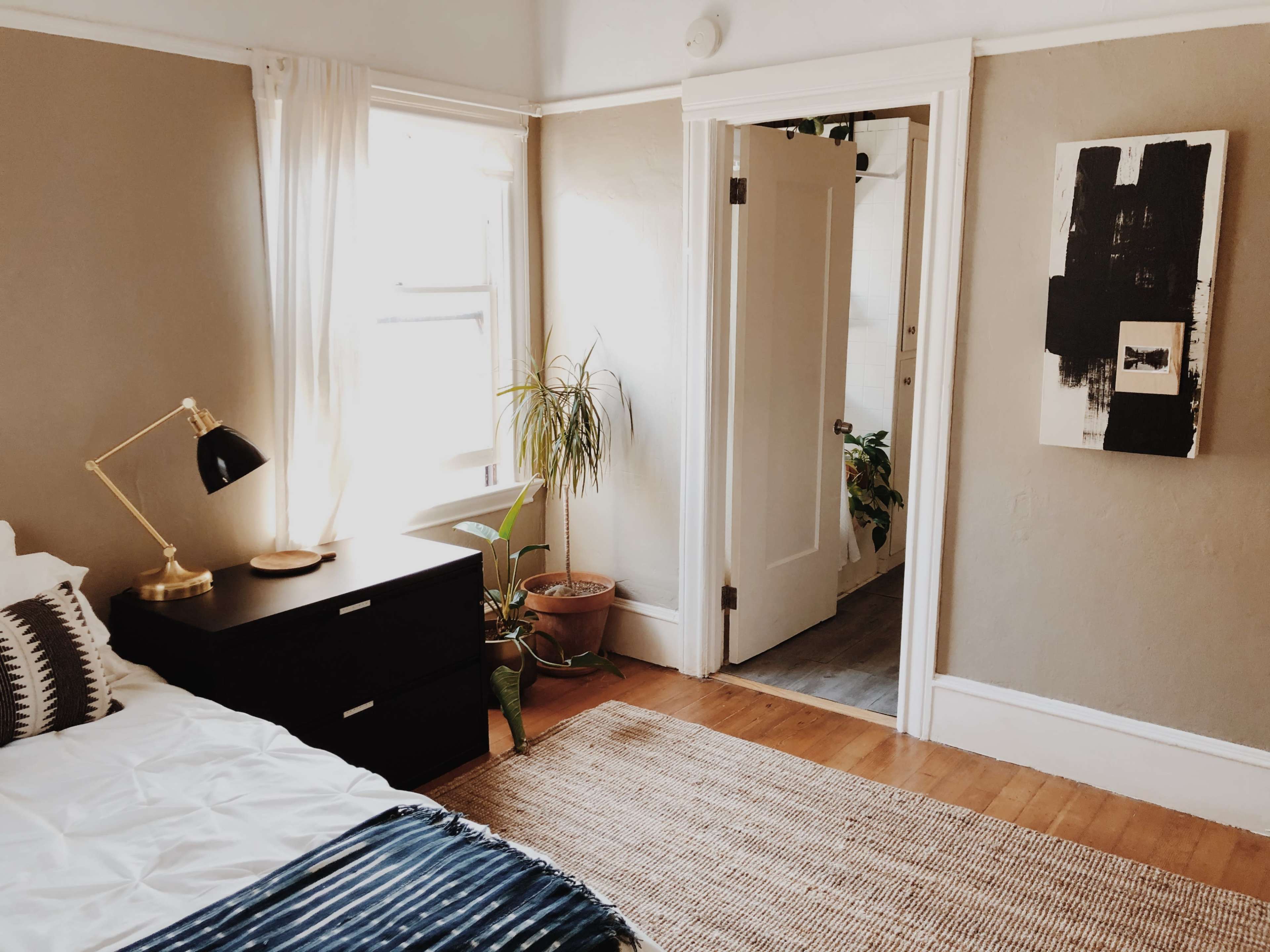 A well-lit bedroom features a bed with decorative pillows, a nightstand with a lamp, and a doorway leading to a hallway with plants.