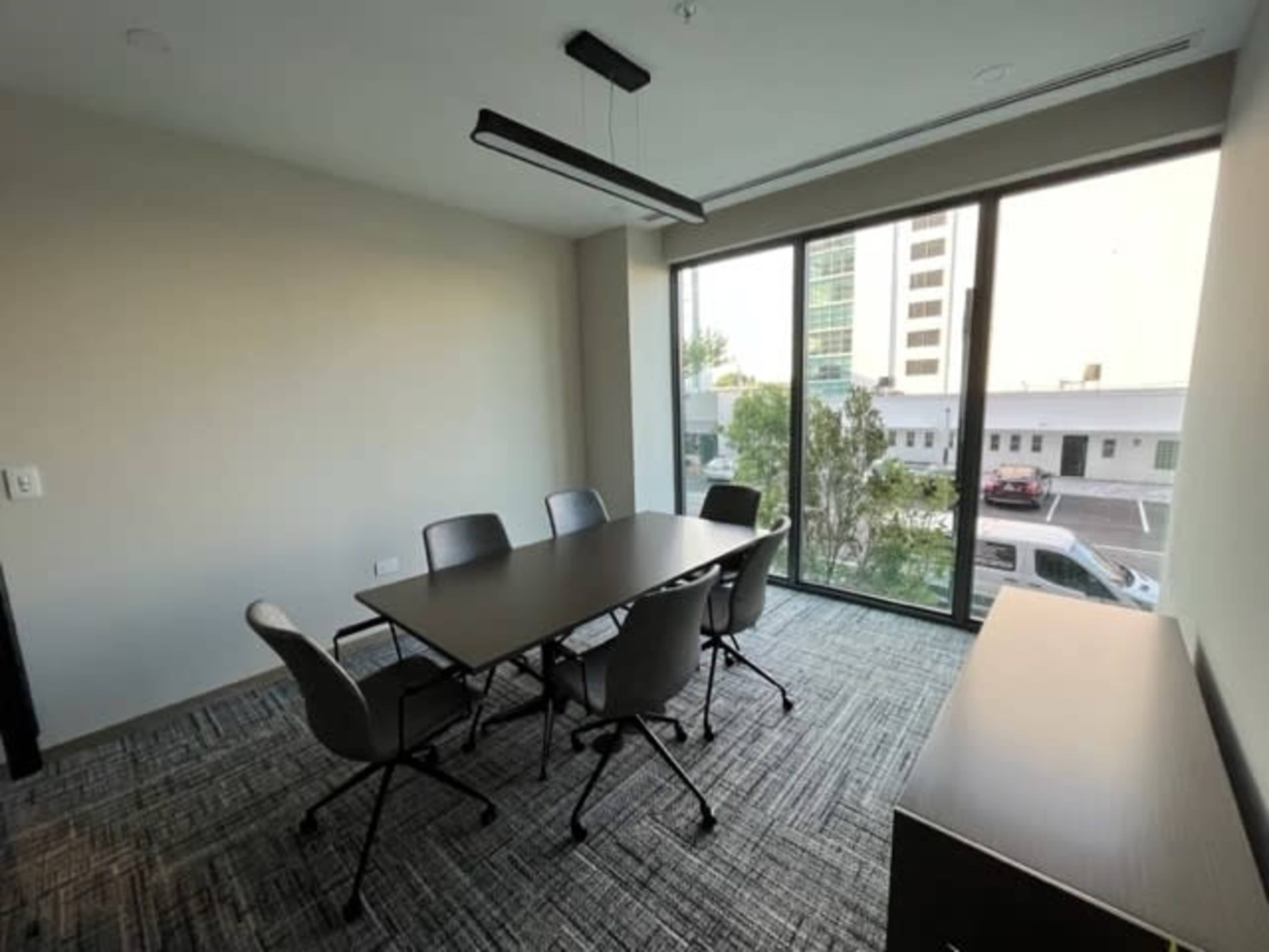 A conference room features a rectangular table surrounded by five chairs, with large windows providing a view of an outside area.