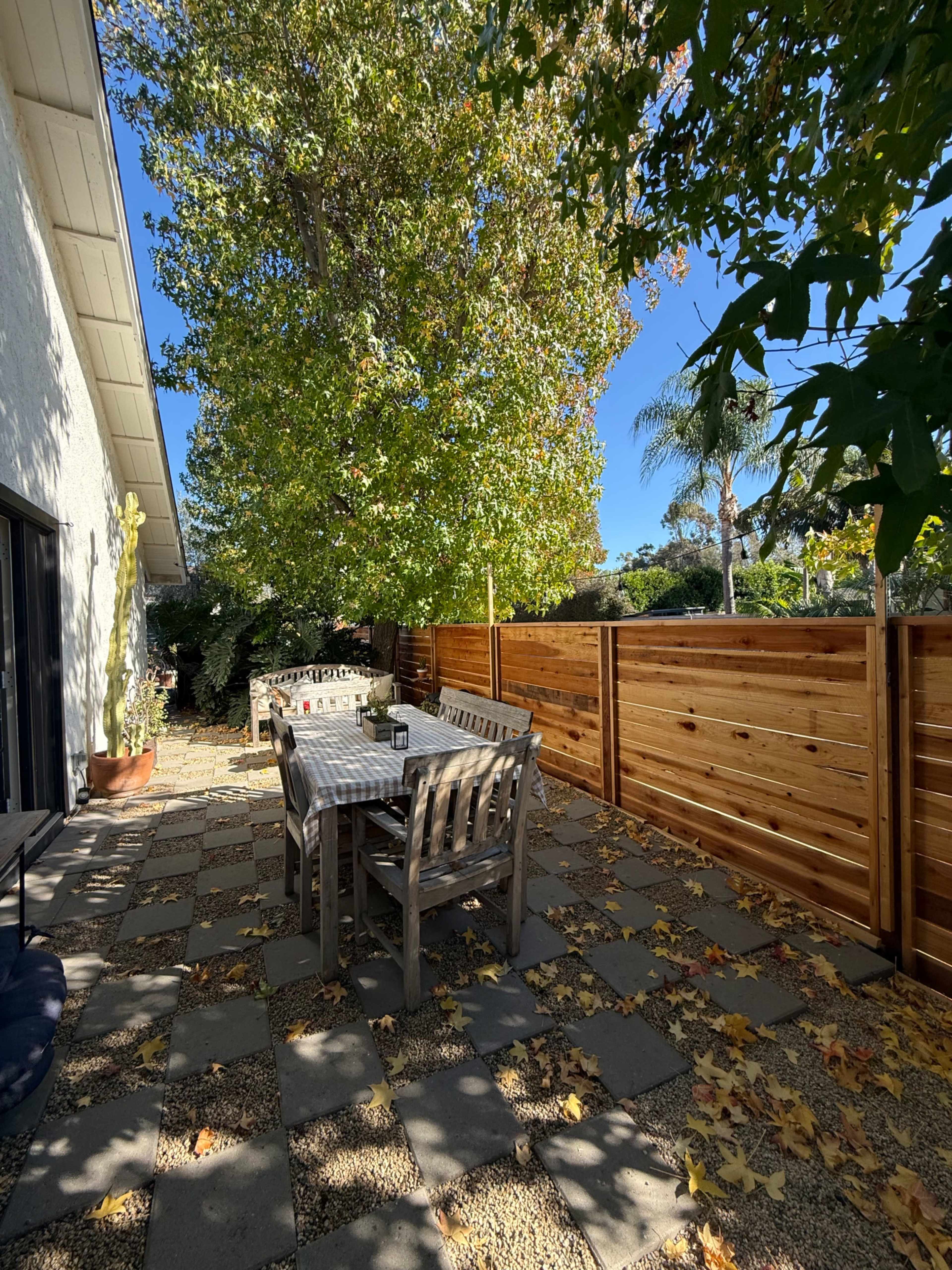 A wooden dining table with chairs is set on a stone patio surrounded by falling leaves, near a fence and a large tree under a clear blue sky.