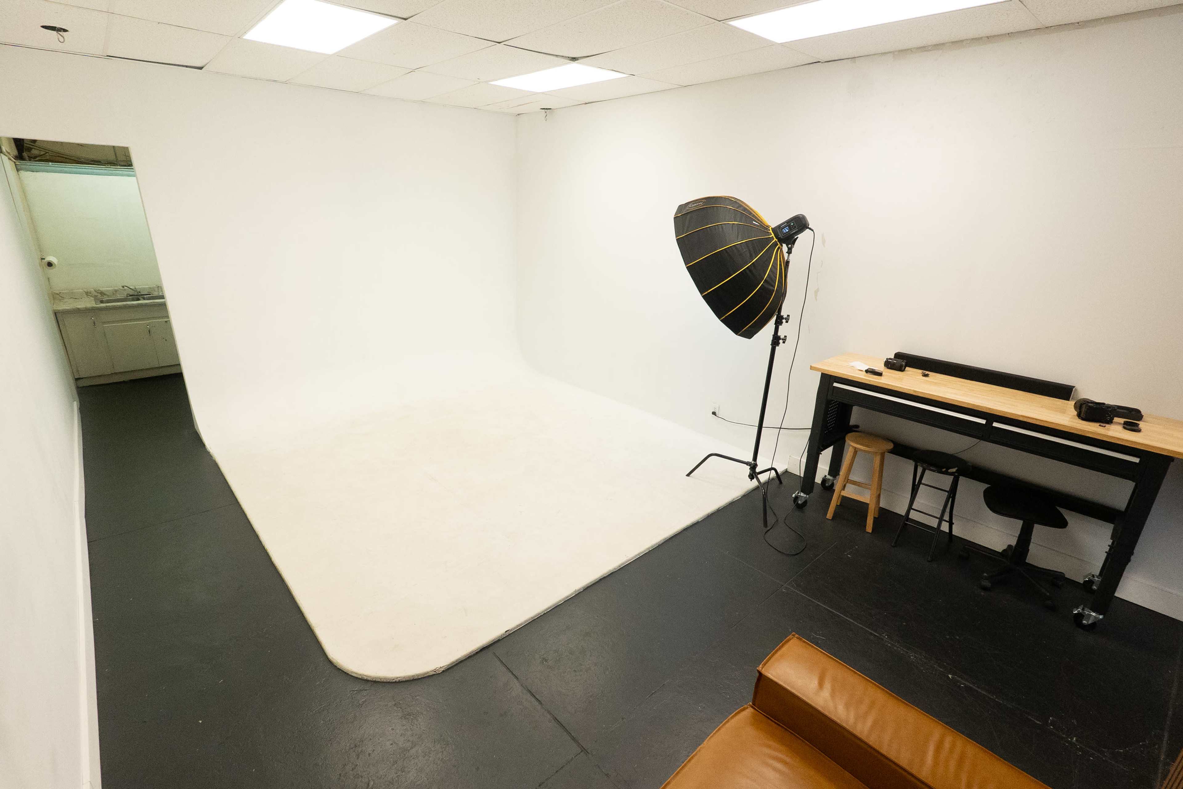 The image shows a minimalist photography studio featuring a white backdrop, a large light source, and a wooden table with chairs in one corner.