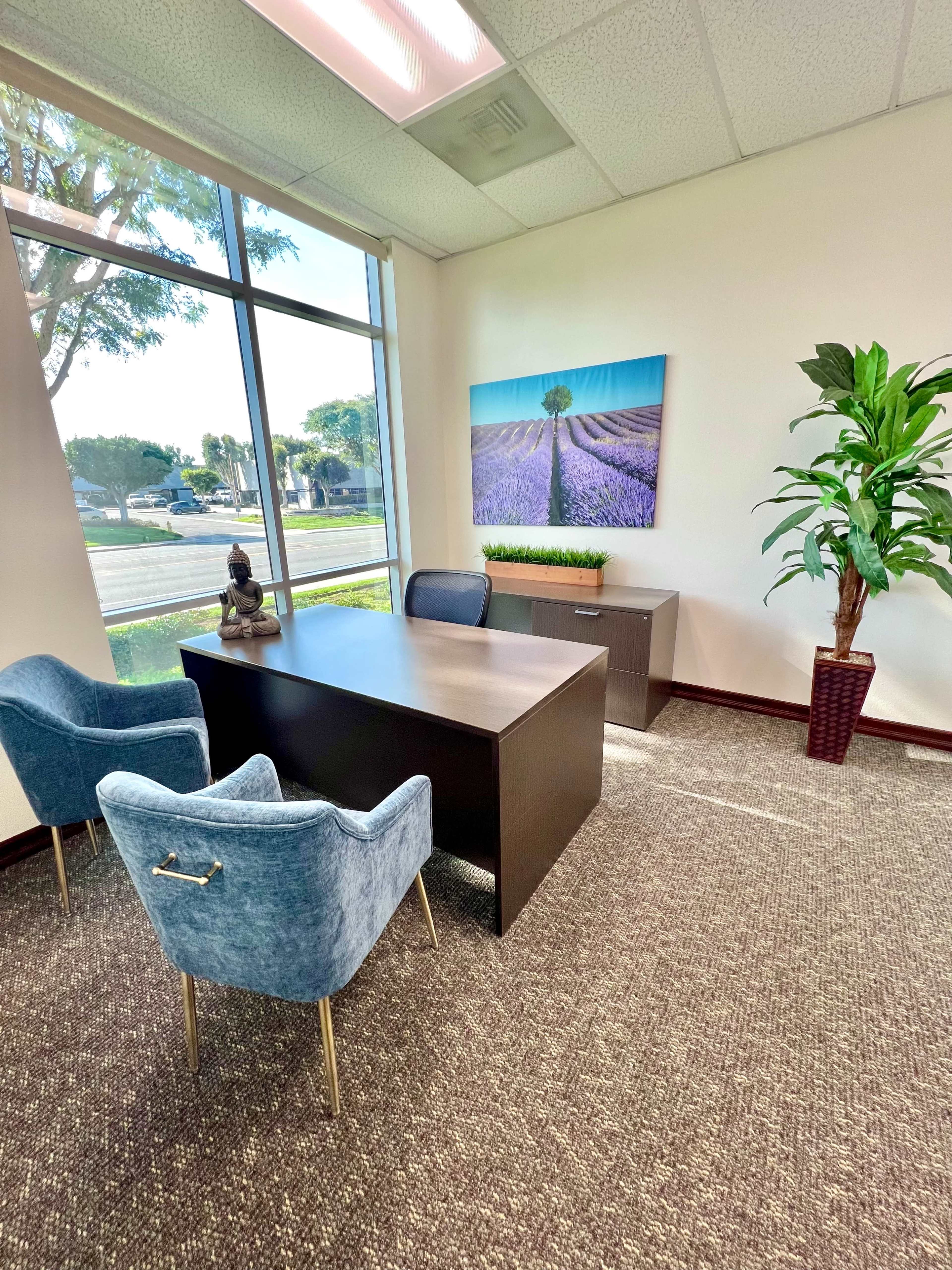 The image shows a modern office with a desk, two blue chairs, a plant, and a large wall art depicting a tree in a lavender field, all illuminated by natural light from large windows.