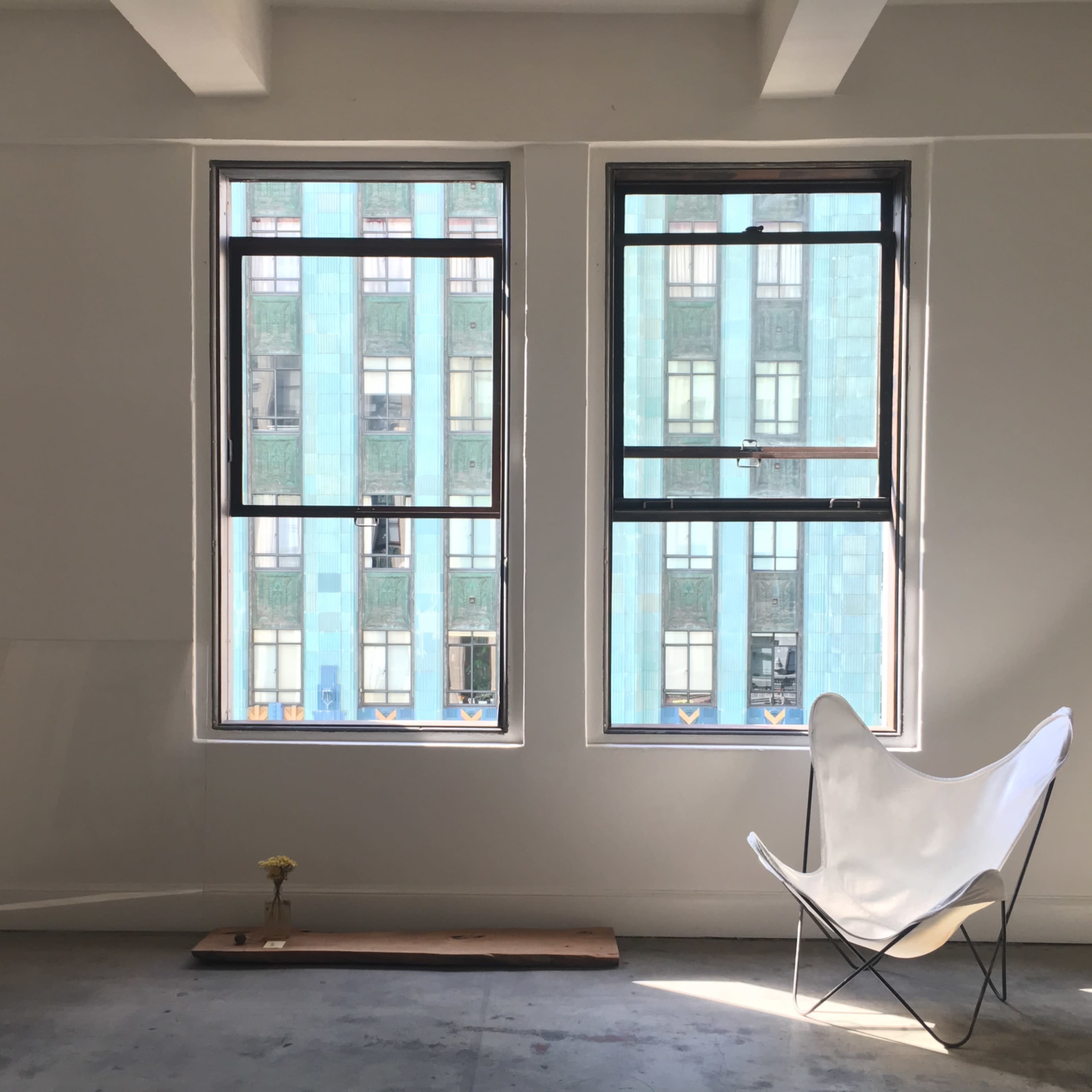 A simple interior space features two large windows framing a view of a blue building, with a butterfly chair and a small flower arrangement on the floor.