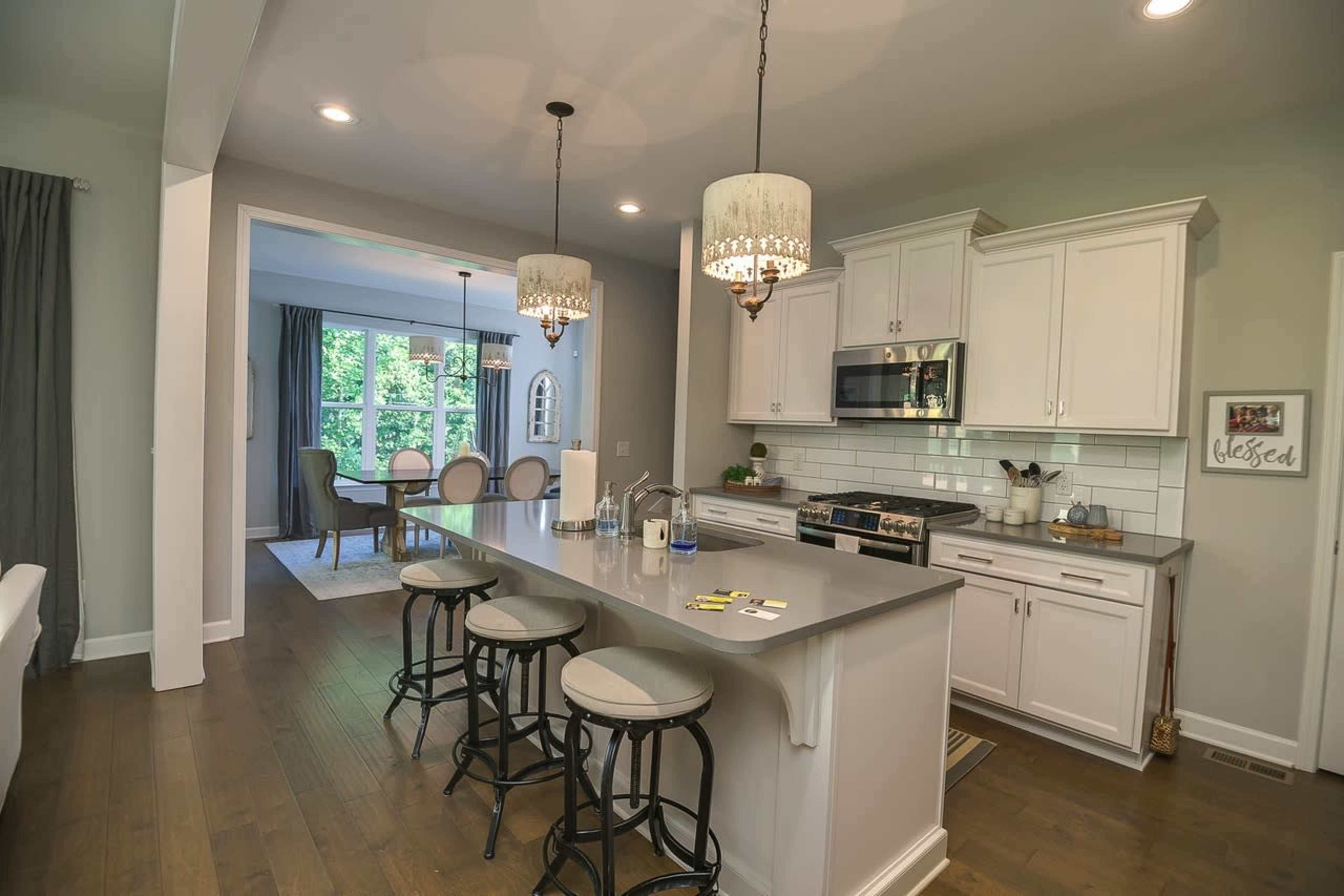 The image shows a modern kitchen with white cabinets, stainless steel appliances, and a large island featuring three bar stools, connected to a dining area with a table and chairs in the background.