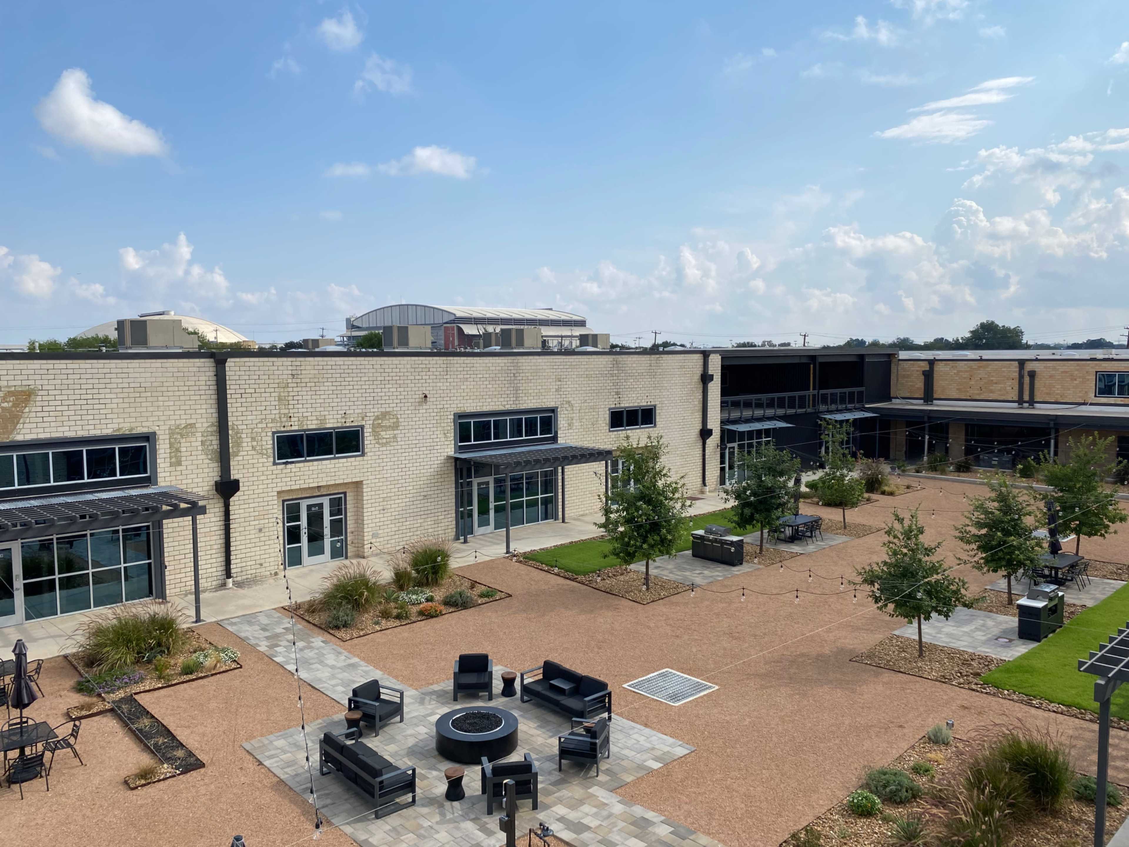 The image shows a modern courtyard surrounded by brick buildings, featuring landscaped areas, seating arrangements, and a fire pit.