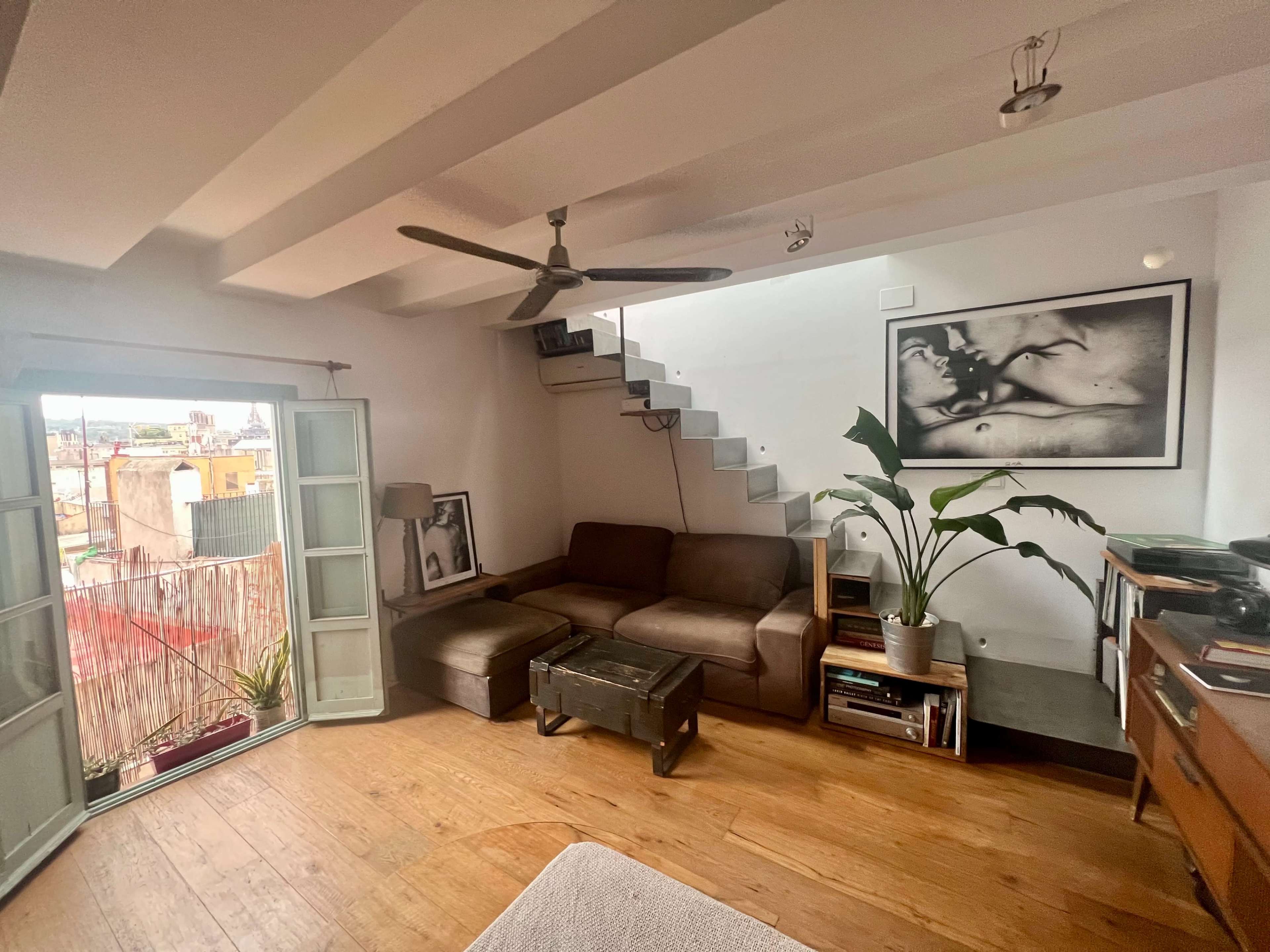 The image shows a cozy living room with a sofa, a coffee table, a staircase leading to an upper level, and a large window allowing natural light to enter.