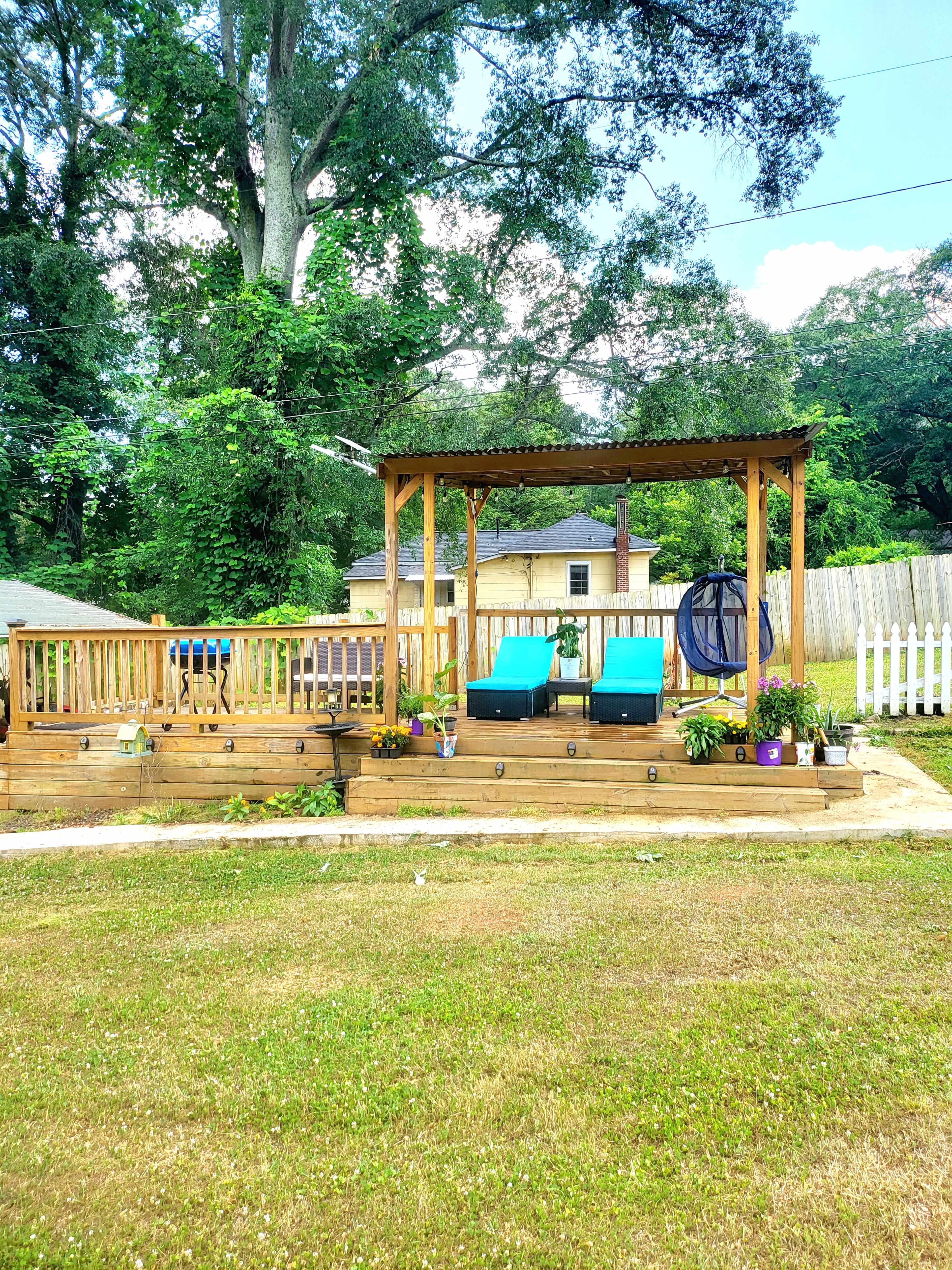 A wooden deck with two teal lounge chairs under a pergola, surrounded by potted plants and a grassy area.