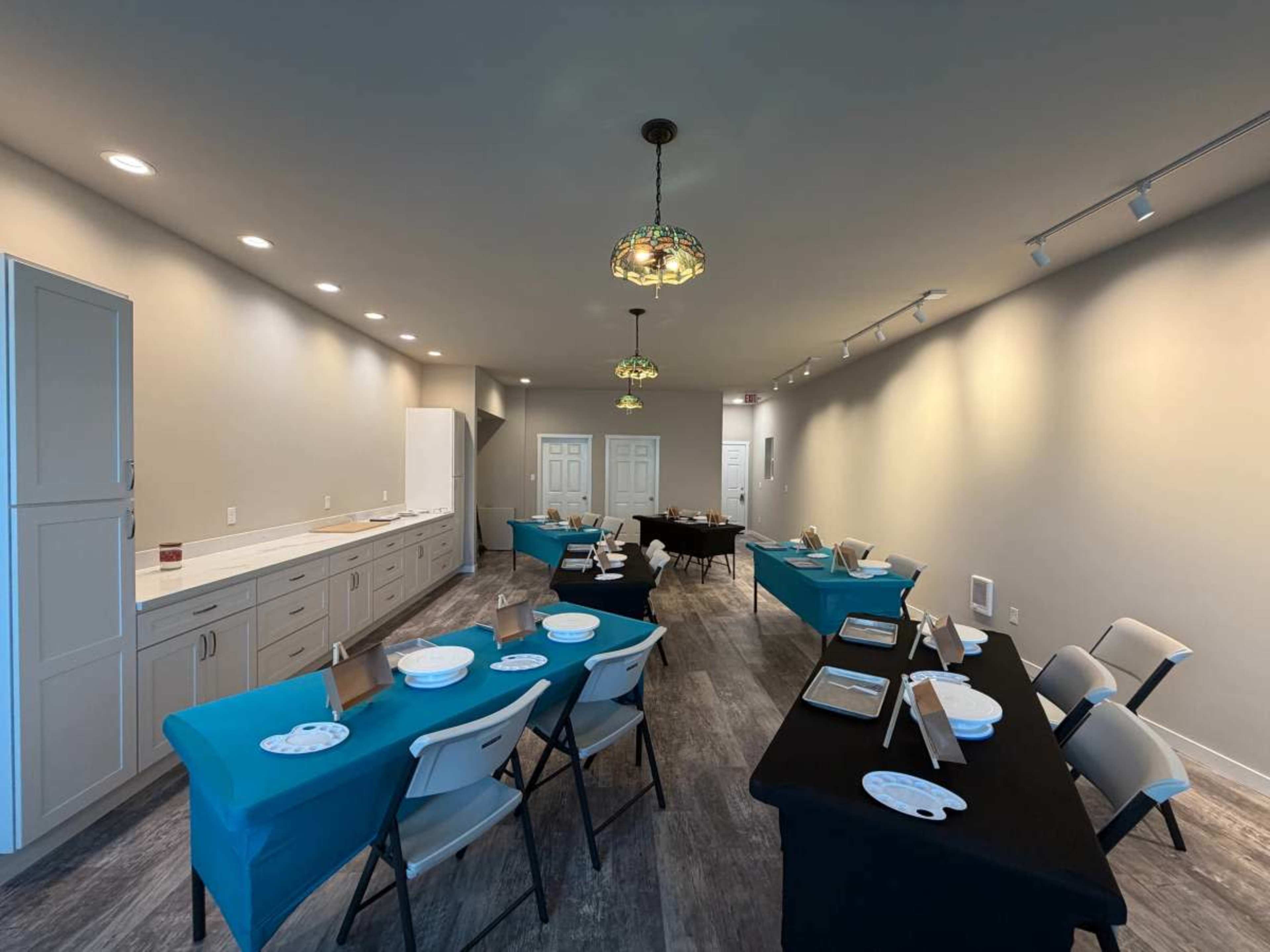 The image shows a modern meeting room with several tables set up, some covered with black cloth and others with teal, along with chairs arranged around them and vintage-style light fixtures hanging from the ceiling.