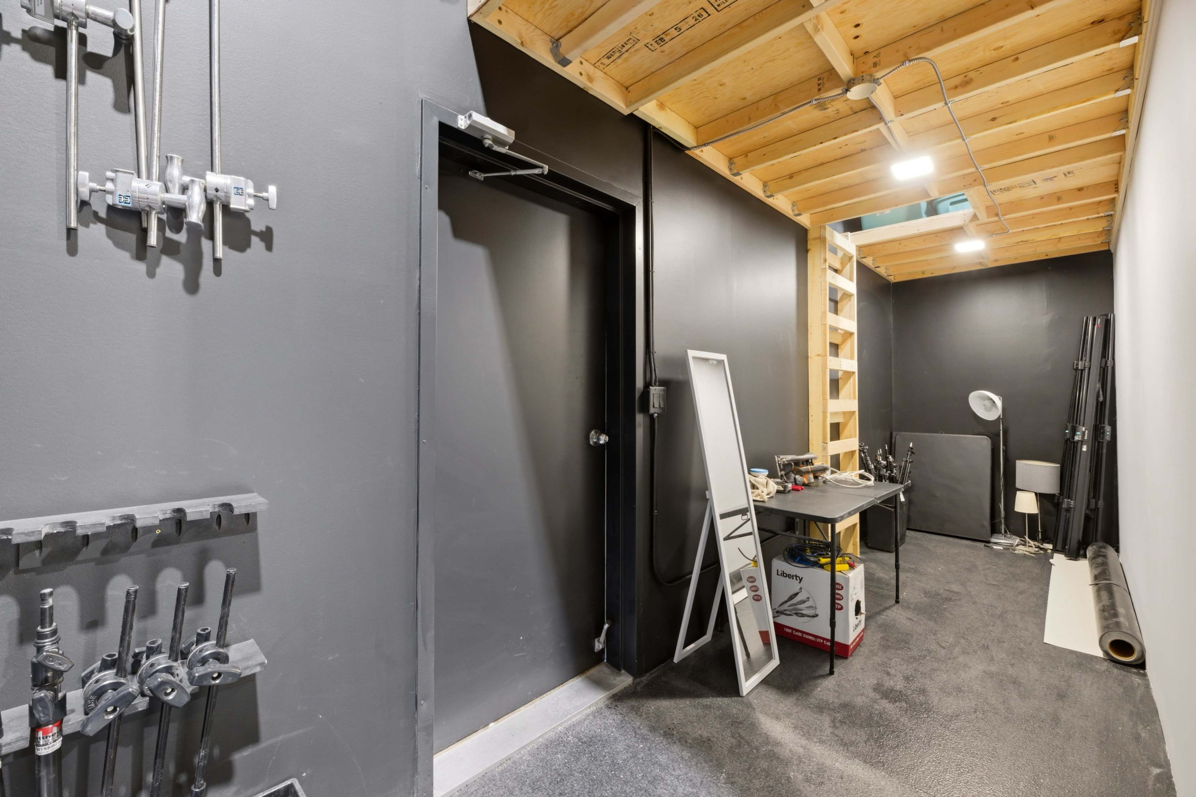 The image shows a narrow hallway with a black wall, a door on one side, and shelves on the opposite side, alongside a table with various tools and equipment.