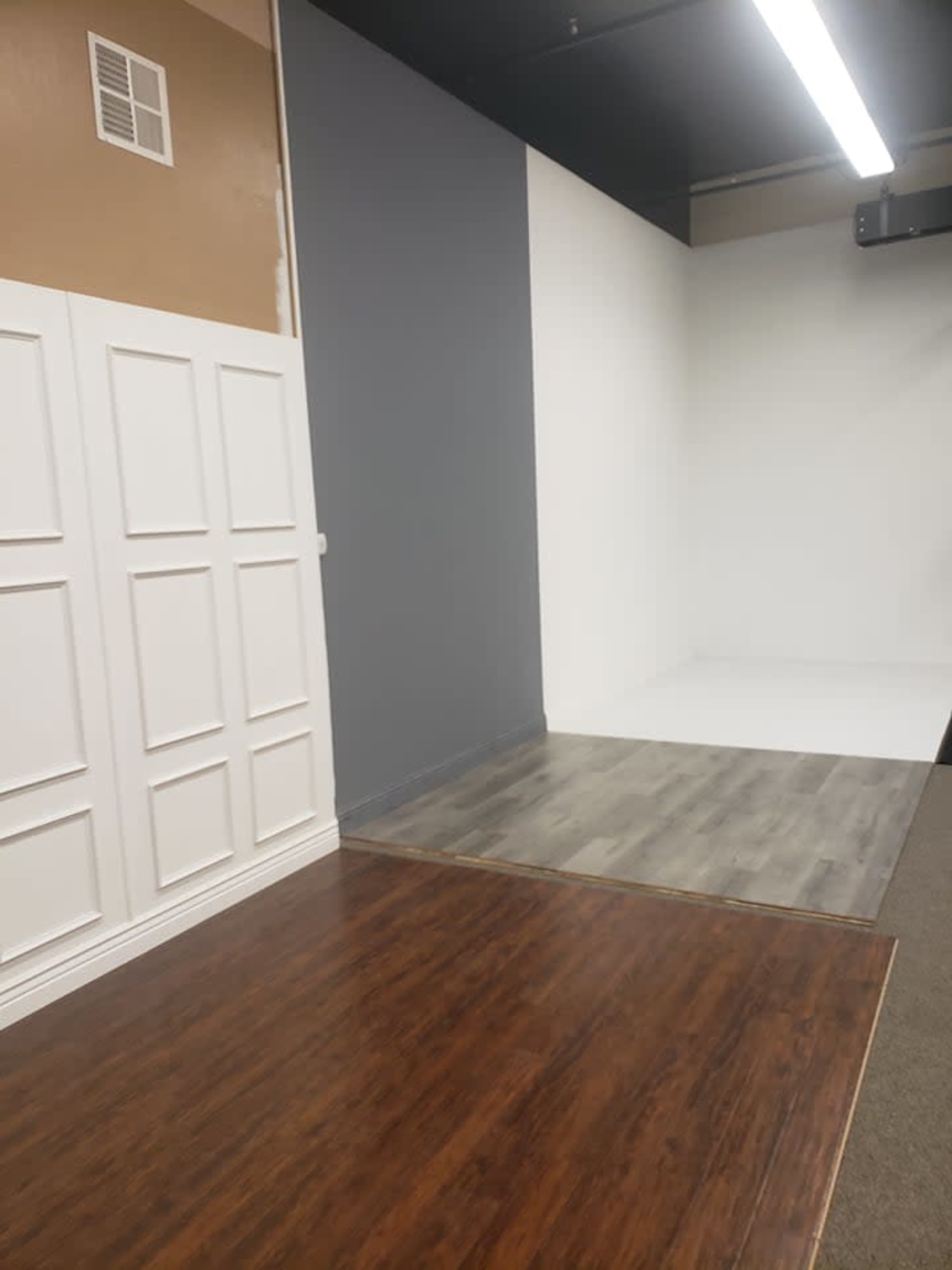 A room featuring three distinct wall sections: one with a white paneled design, one painted gray, and one painted white, with different flooring materials visible.