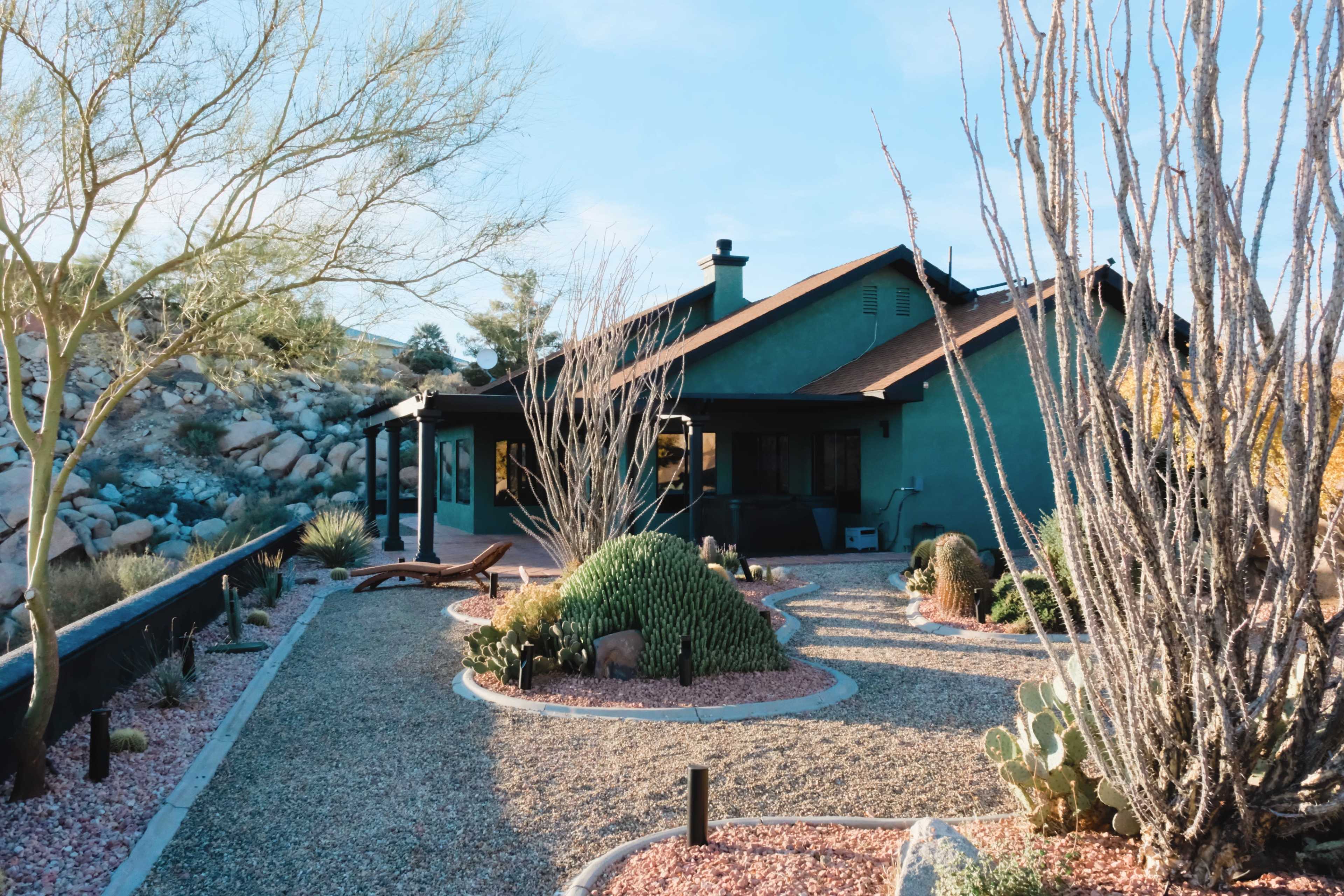 A green house with a sloped roof is surrounded by gravel landscaping and desert plants under a clear sky.