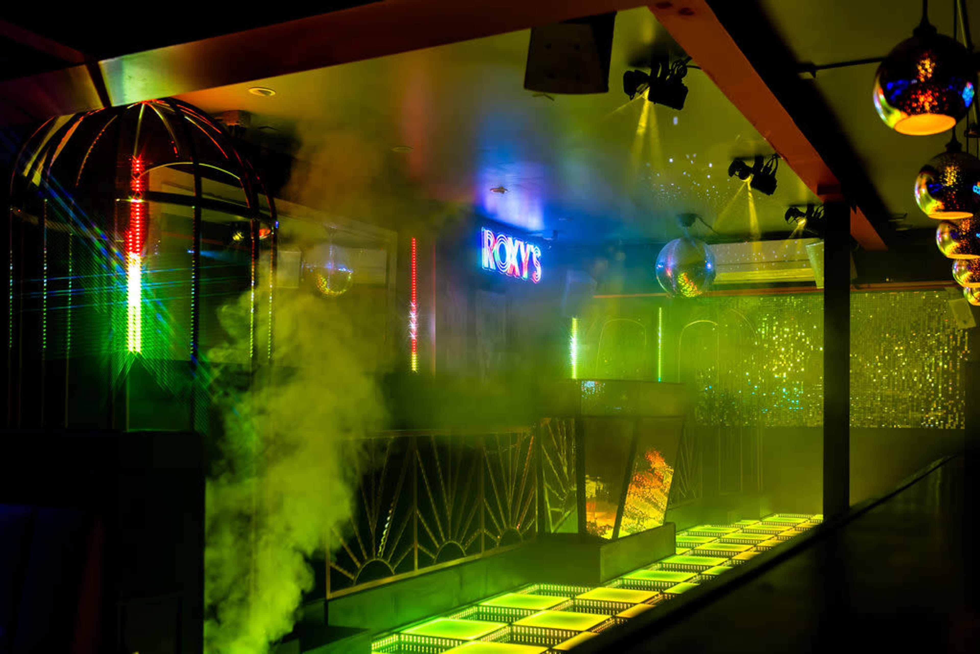 The image shows a vibrant nightclub interior with colorful lighting, disco balls, and a misty atmosphere.