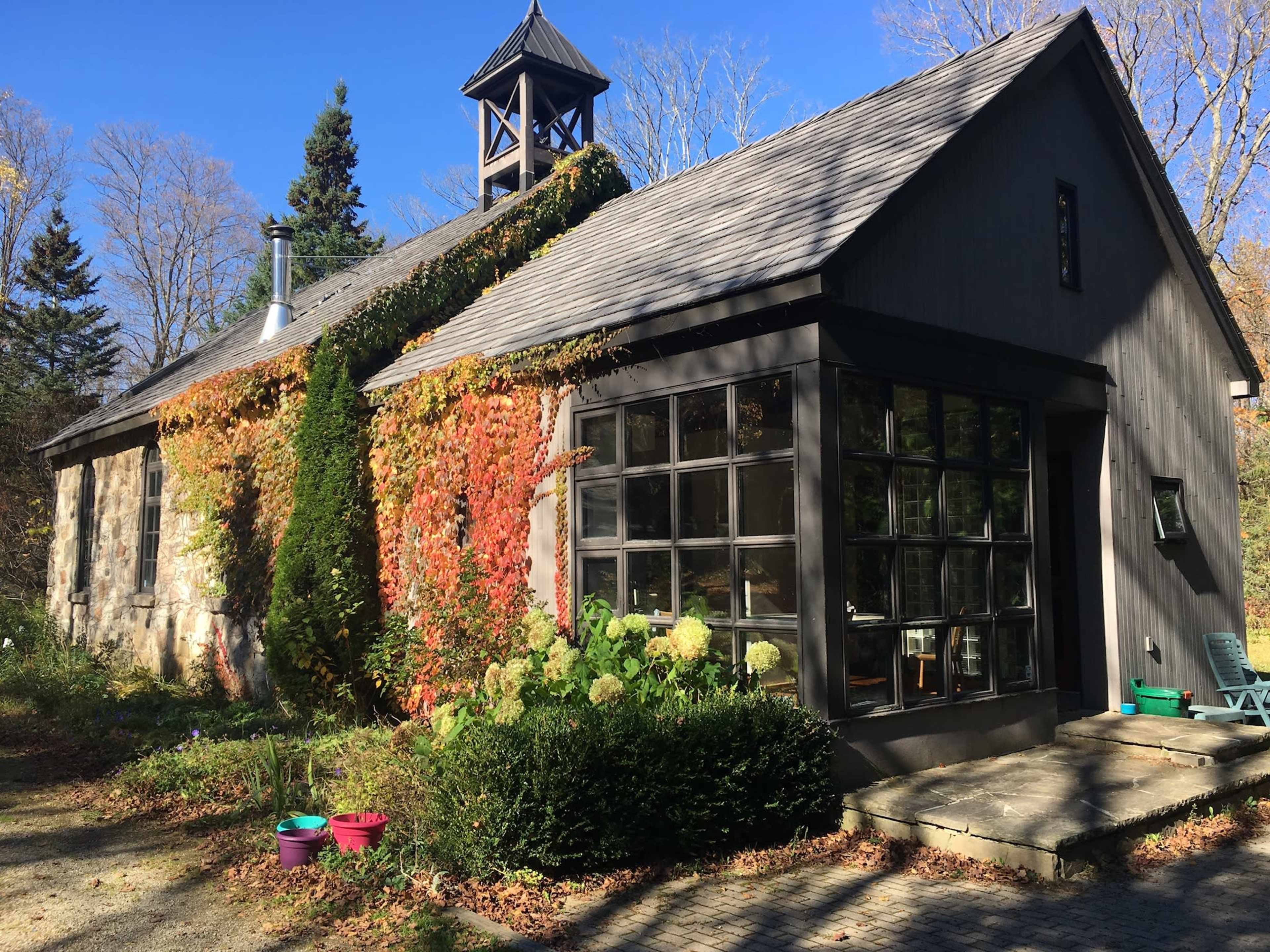 A stone and wooden house features a large glass wall, surrounded by greenery and colorful foliage.