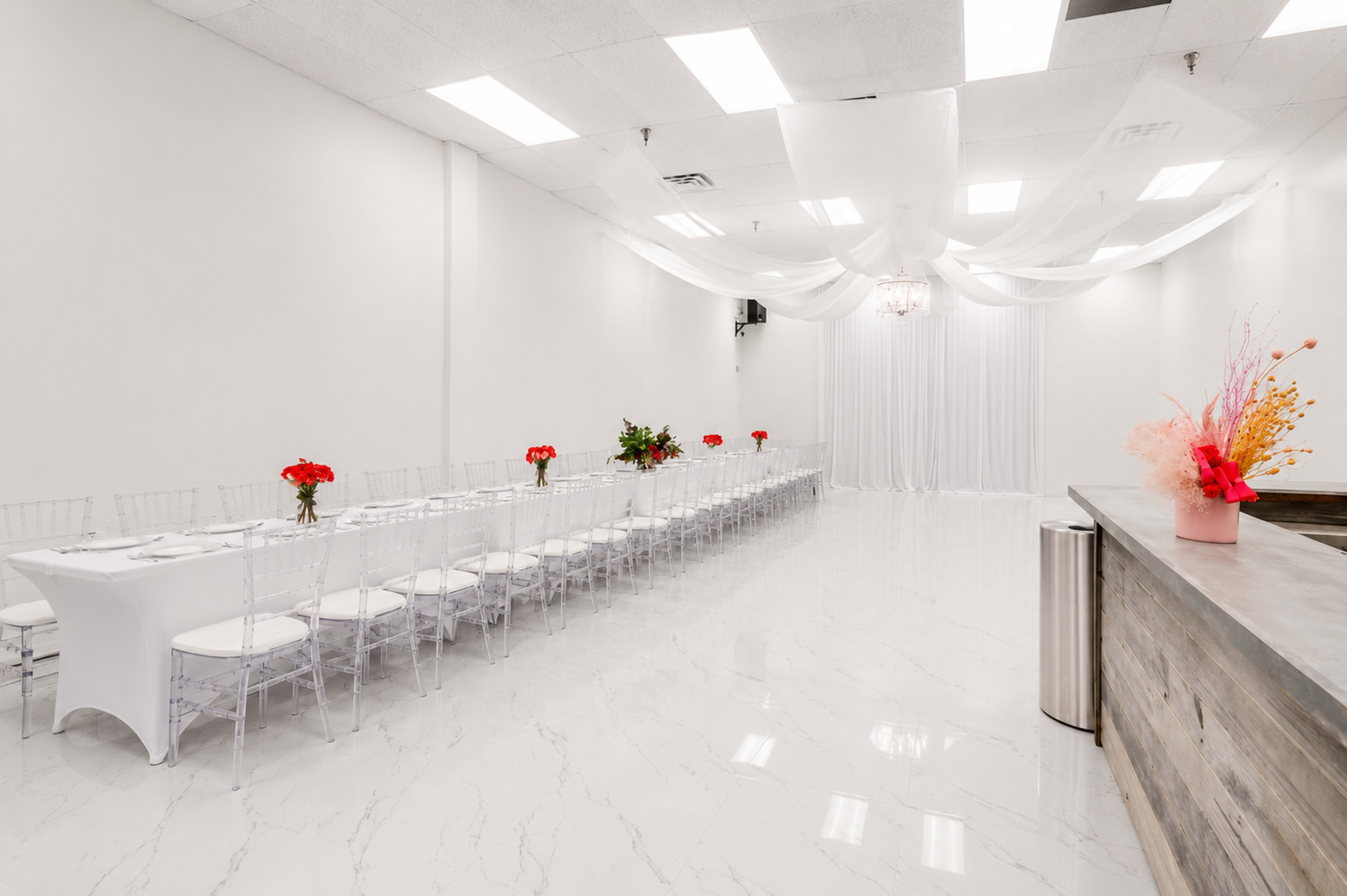 The image shows a banquet hall with a long dining table set for an event, adorned with red floral centerpieces and surrounded by clear acrylic chairs, all under soft draped fabric overhead.