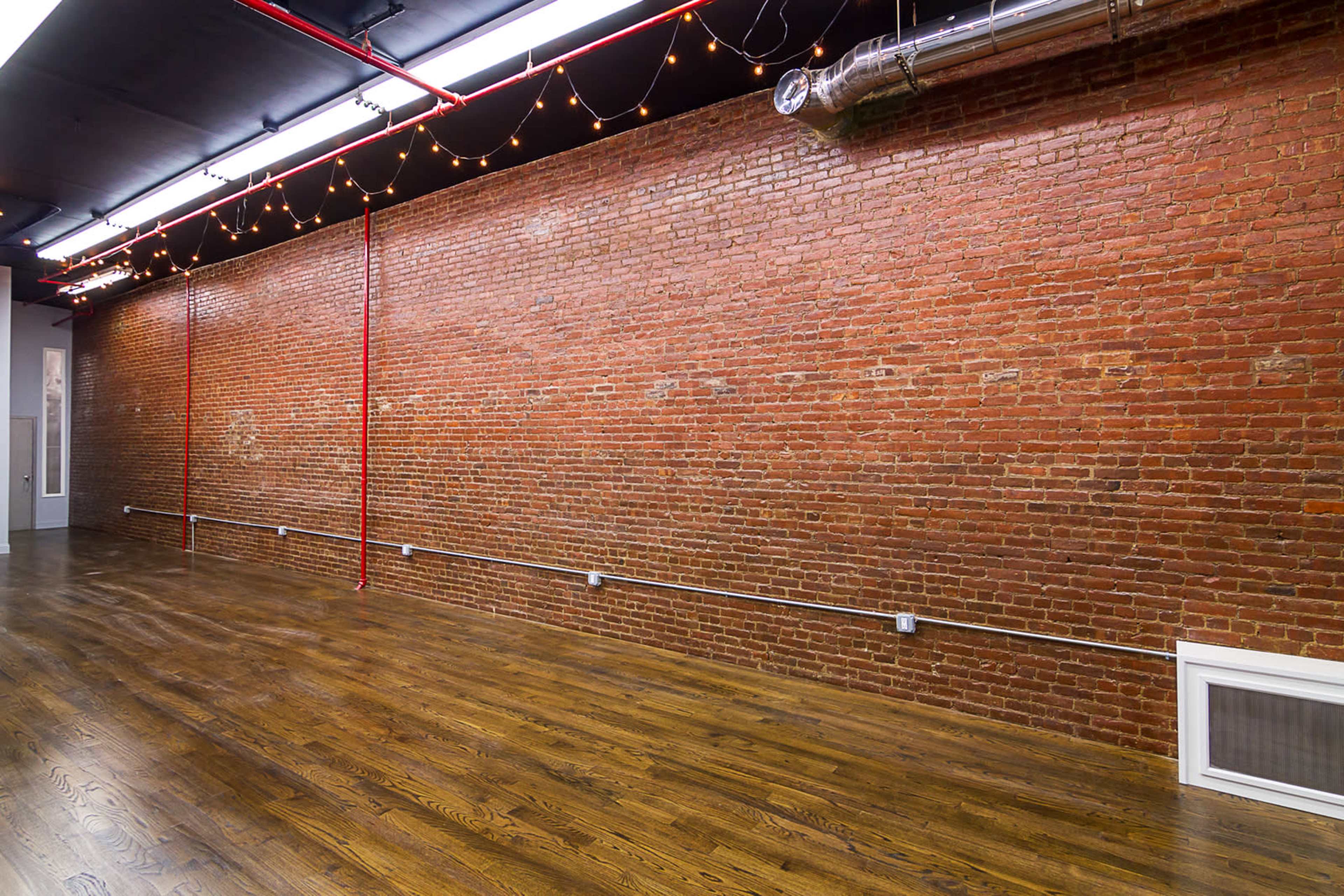 The image shows a spacious interior featuring a red brick wall and polished wooden flooring illuminated by overhead lights and string lights.