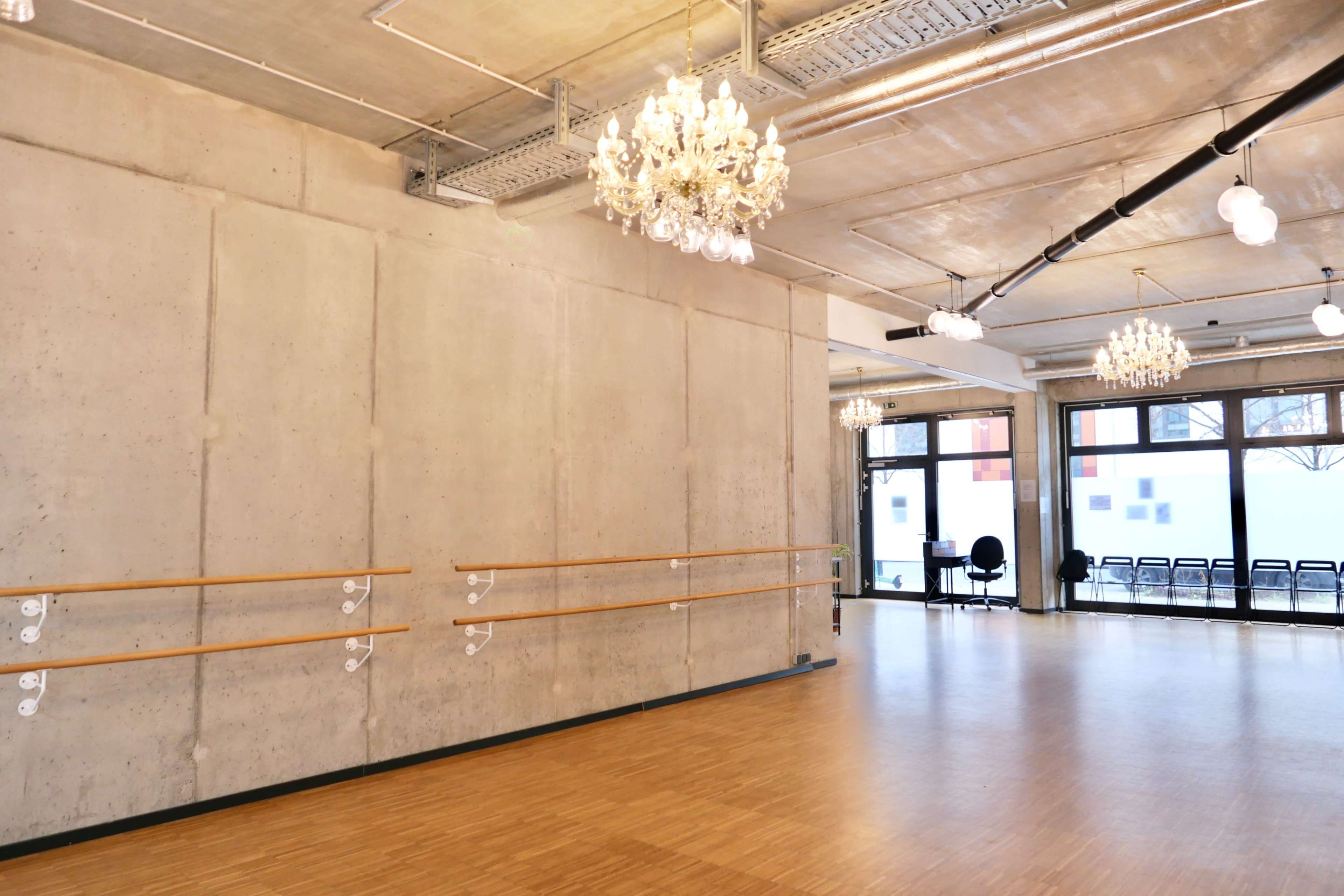 The image shows a spacious dance studio with a wooden floor, concrete walls, and several chandeliers hanging from the ceiling.