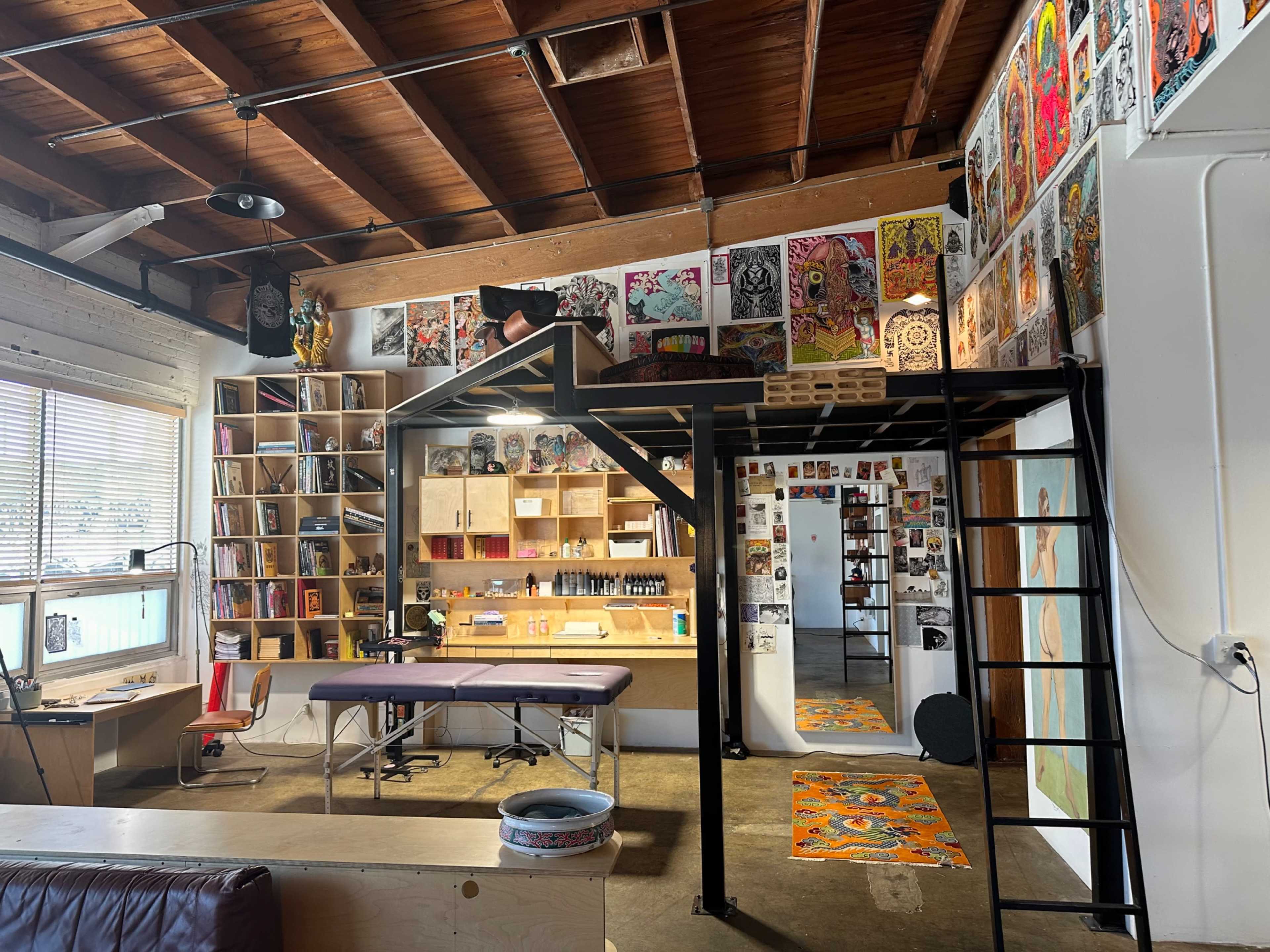 The image shows a spacious artist studio featuring a work area with a large table, shelves filled with art supplies and artworks, and a lofted sleeping area above.