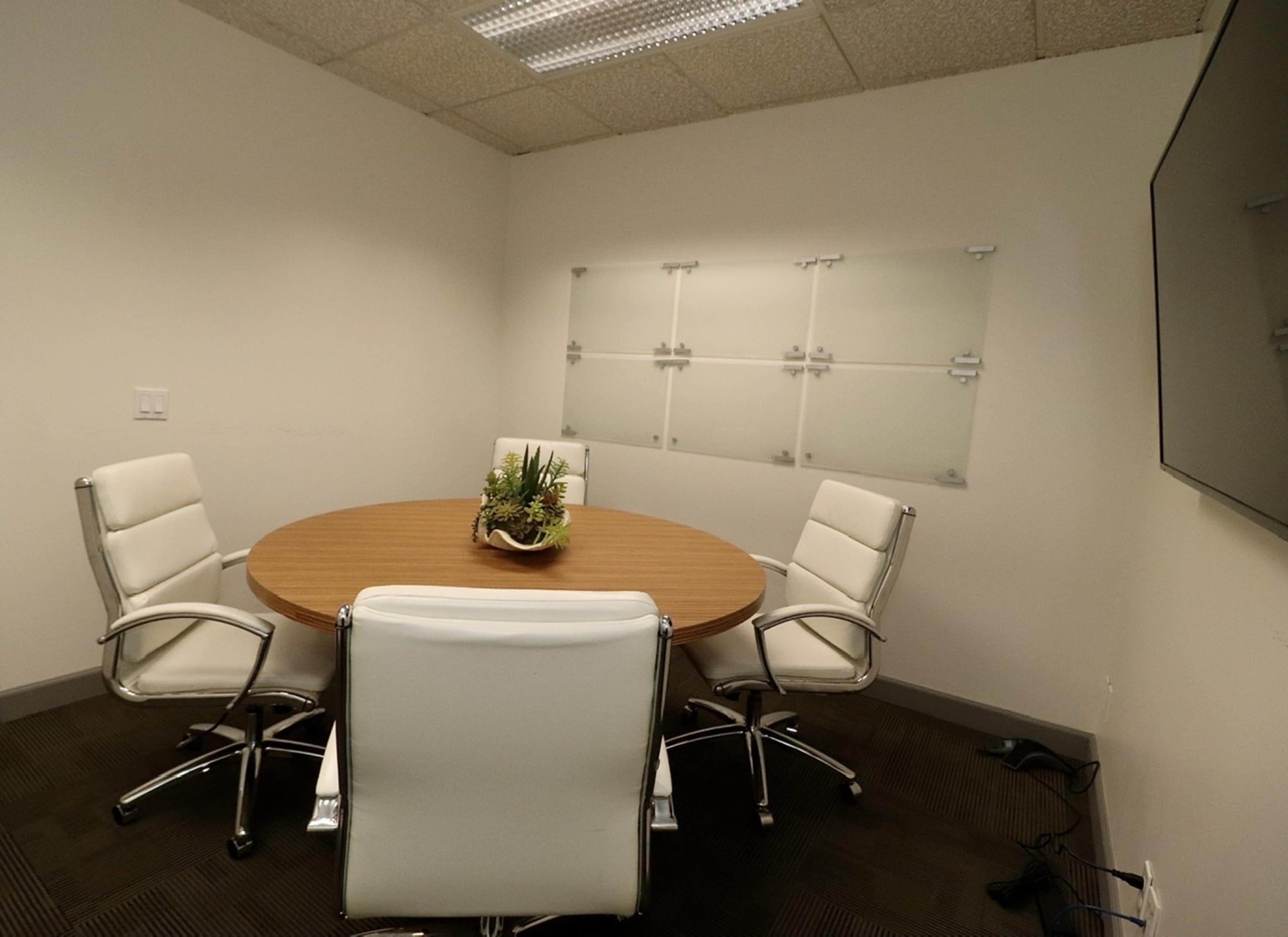 A modern conference room features a round wooden table surrounded by four white swivel chairs, with a wall-mounted television and a glass-panel display.